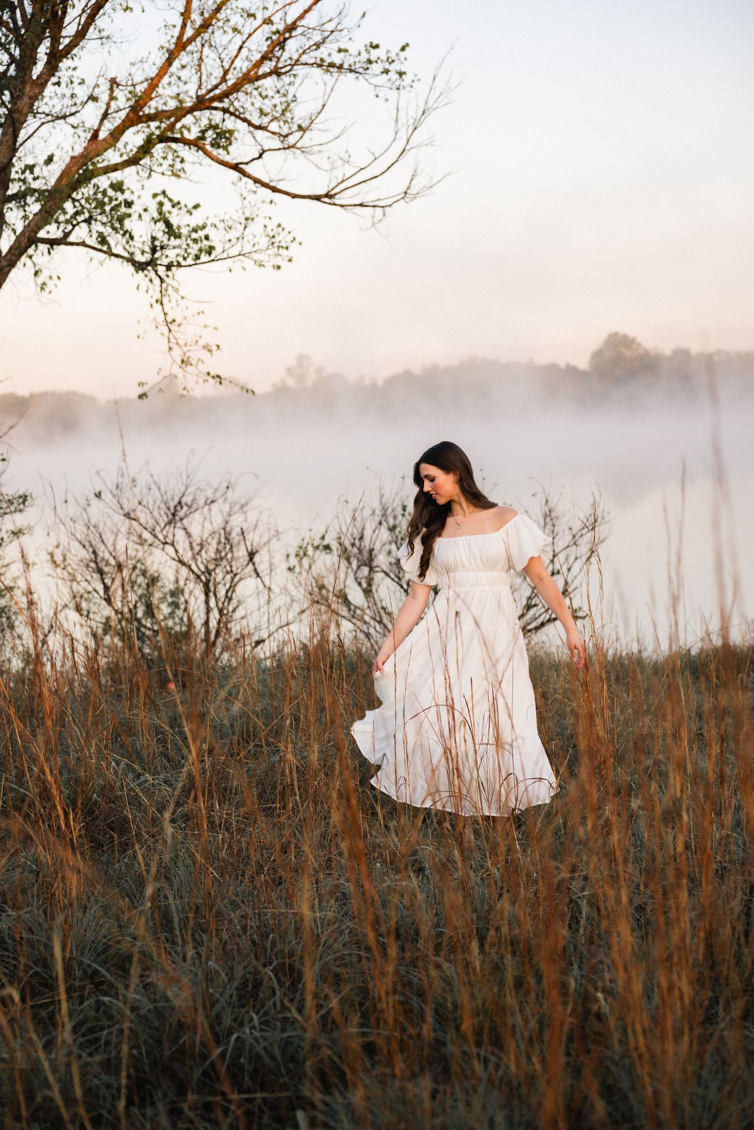 Woman in white off-shoulder dress standing in misty field at golden hour near bare tree.