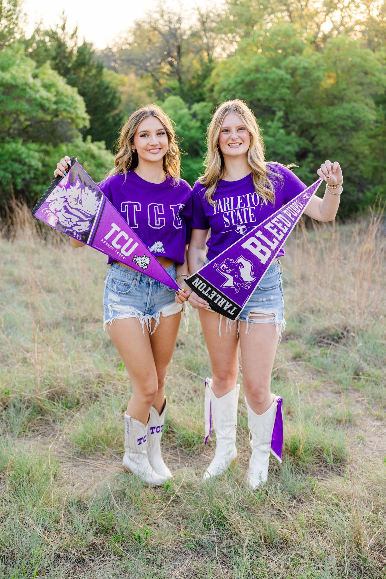 Two young women holding TCU and Tarleton State purple pennants in a field, wearing purple shirts and white cowboy boots.
