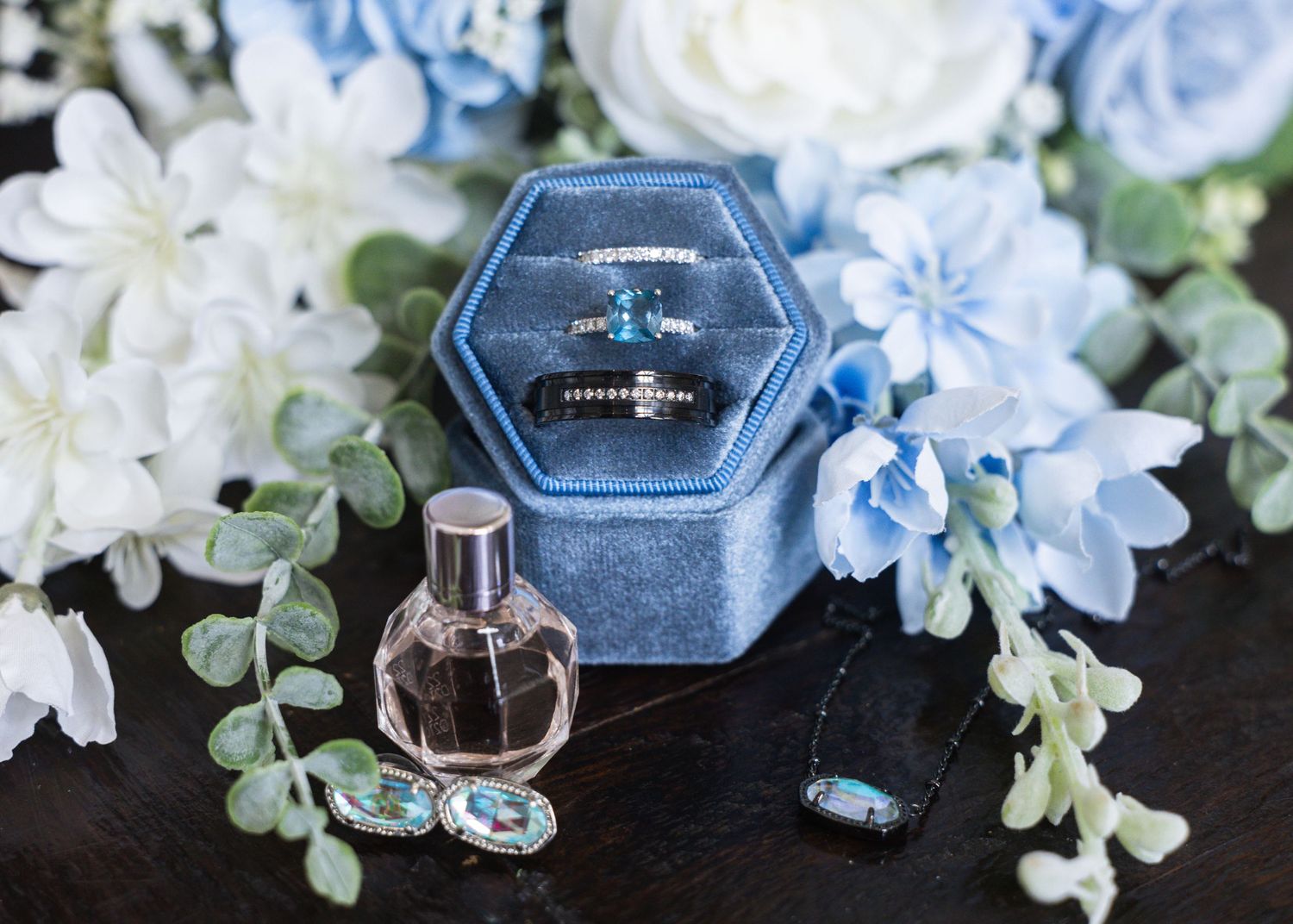Blue velvet hexagon ring box with diamond rings, perfume bottle, and blue and white floral arrangement on dark surface.