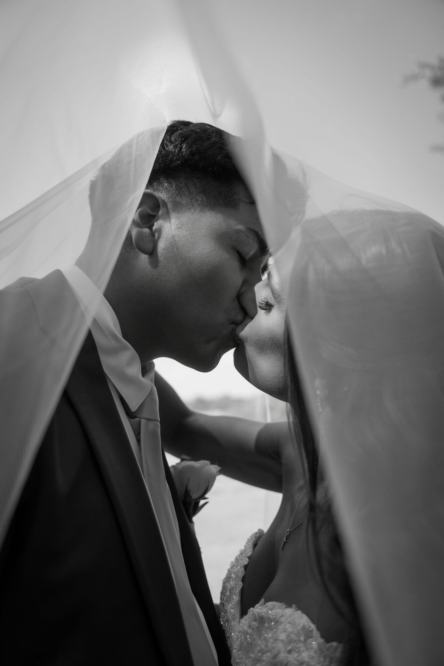 Black and white photo of a bride and groom kissing under a veil on their wedding day.