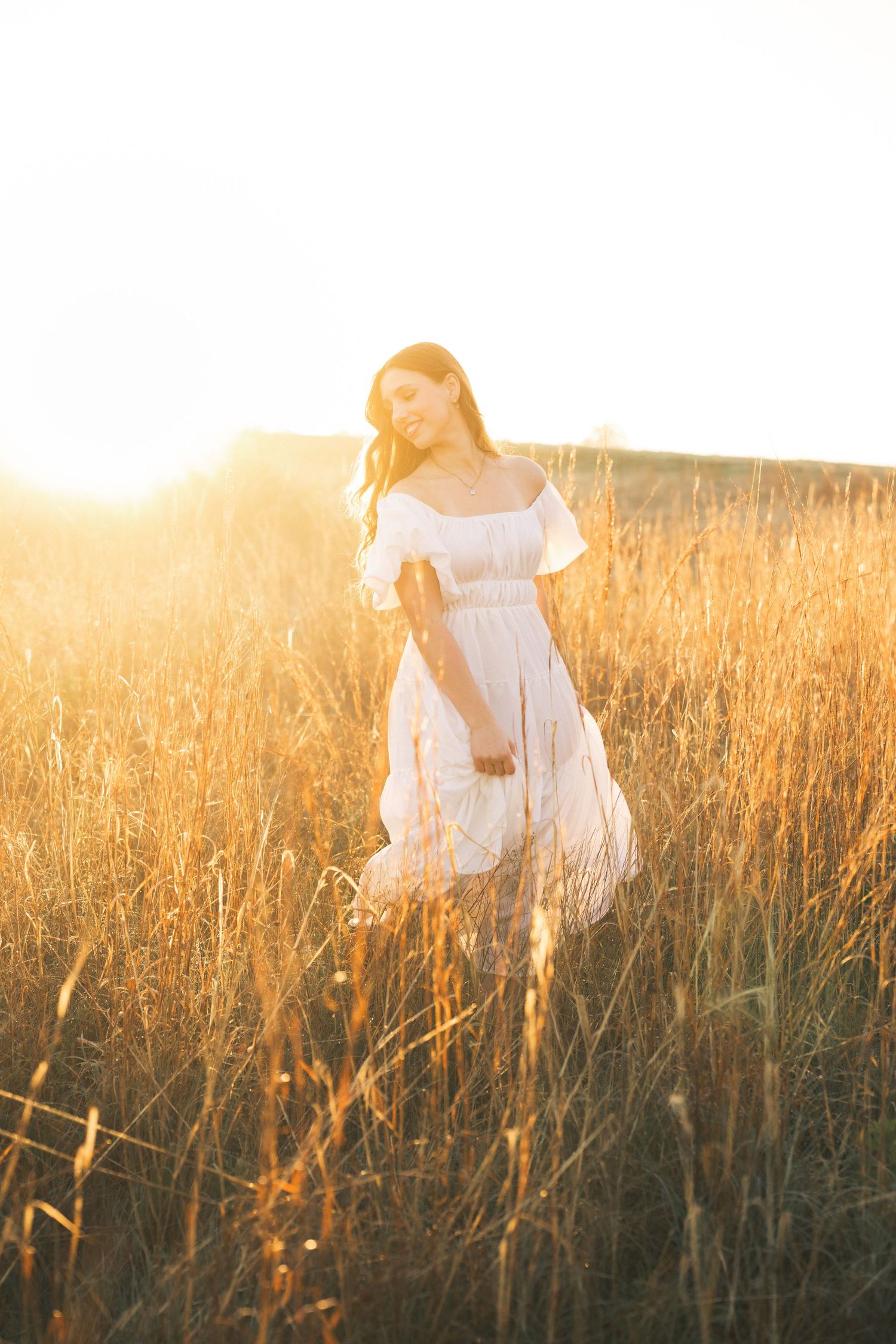 Young woman in white dress standing in golden wheat field at sunset, bathed in warm glowing sunlight.