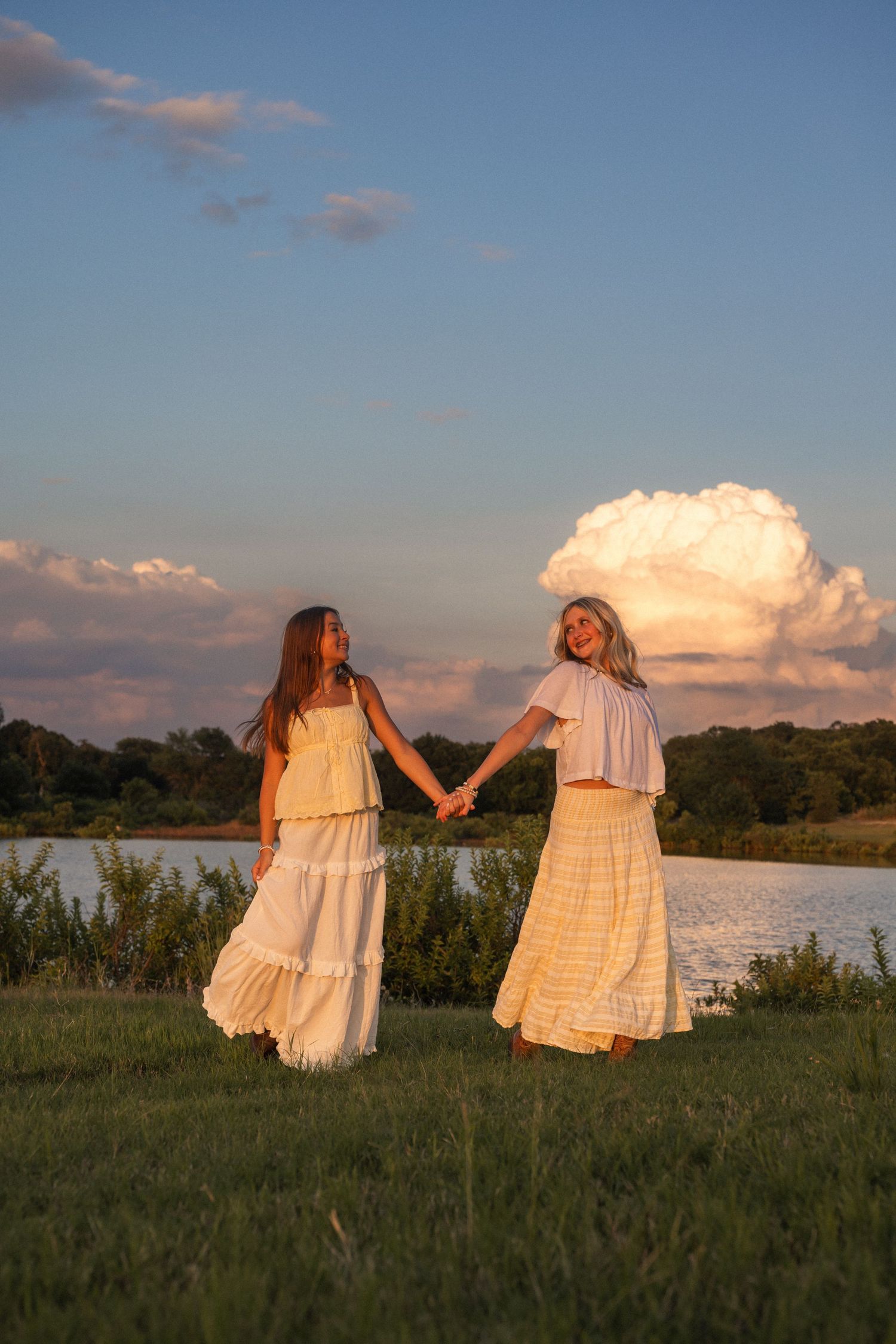 Two girls in white dresses hold hands by a lake at golden hour sunset with dramatic clouds.