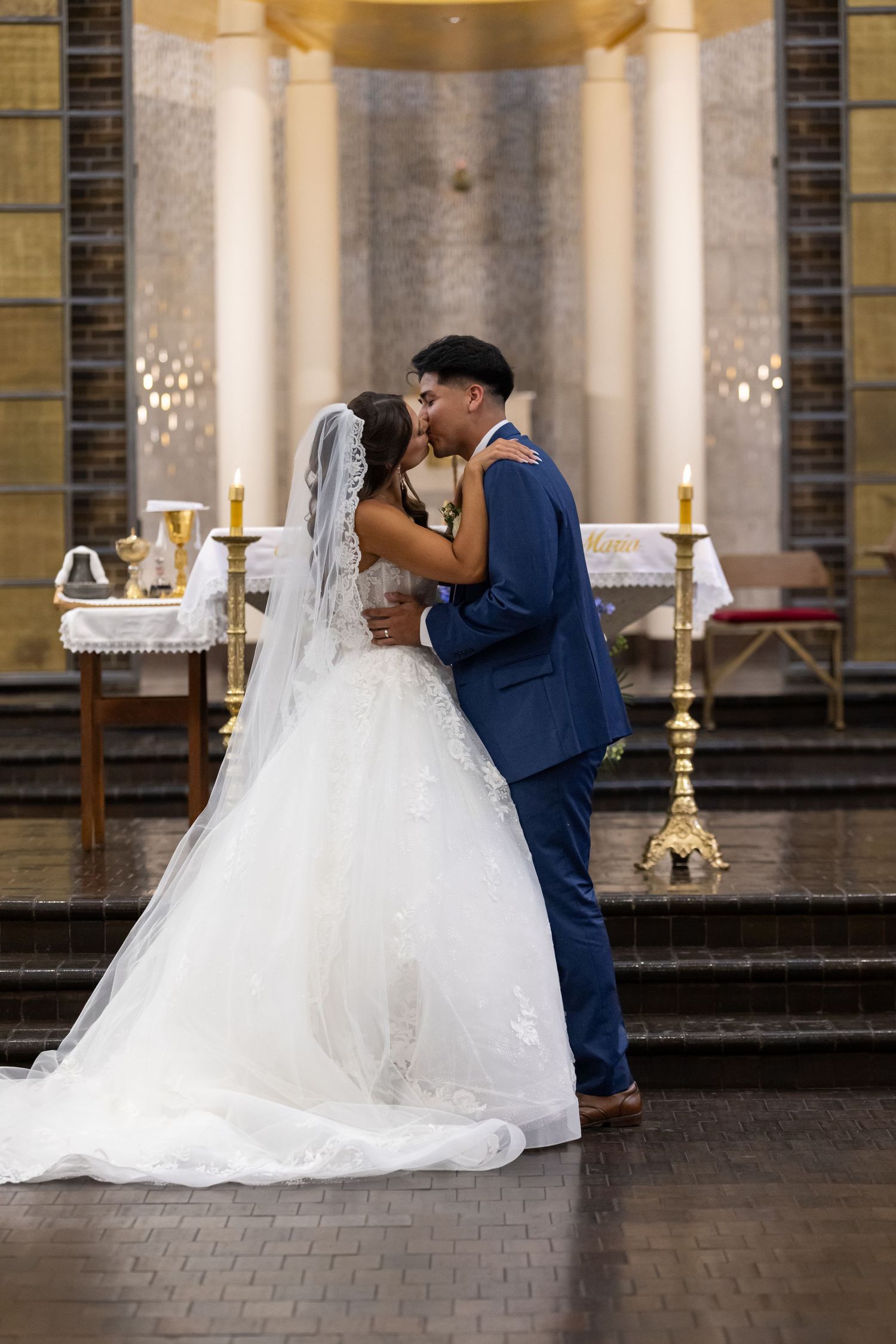 Bride and groom share first kiss at church altar, bride in white gown with veil, groom in navy blue suit.