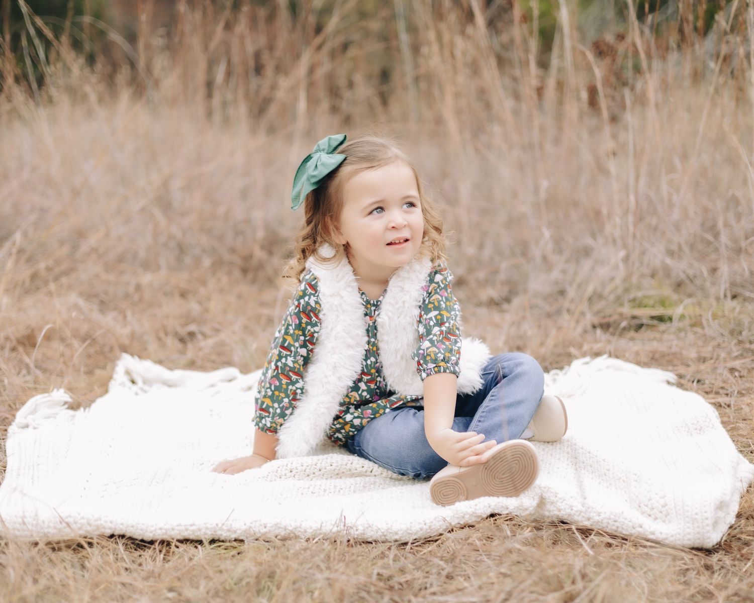 A child in a floral dress and white vest sits on a blanket outdoors in a natural setting.