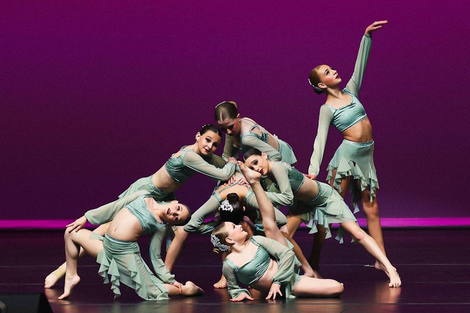 Modern dancers in mint green costumes perform a flowing contemporary dance sequence on a dark purple stage.