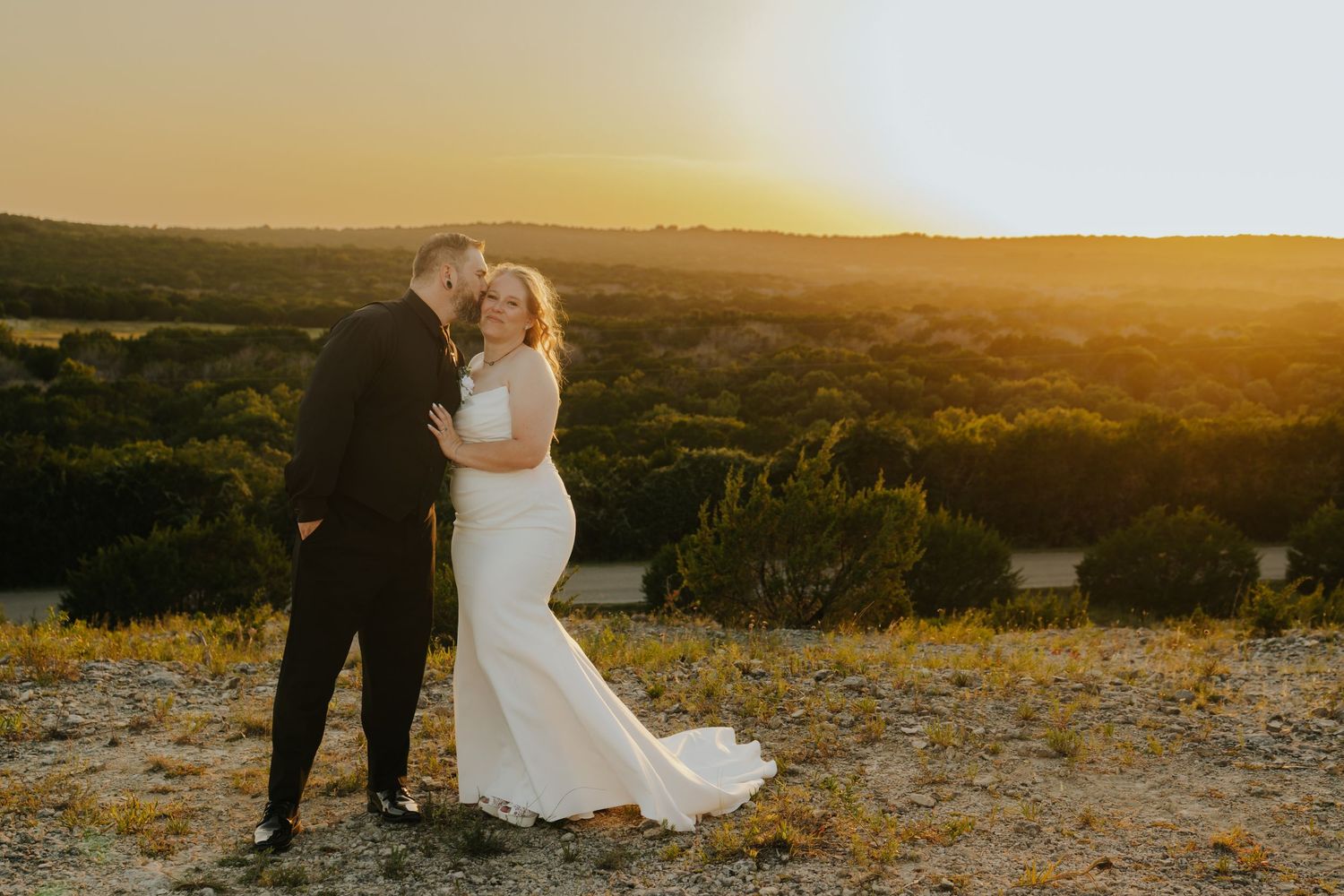 Bride and groom share a romantic sunset kiss overlooking a scenic Texas Hill Country landscape on their wedding day.
