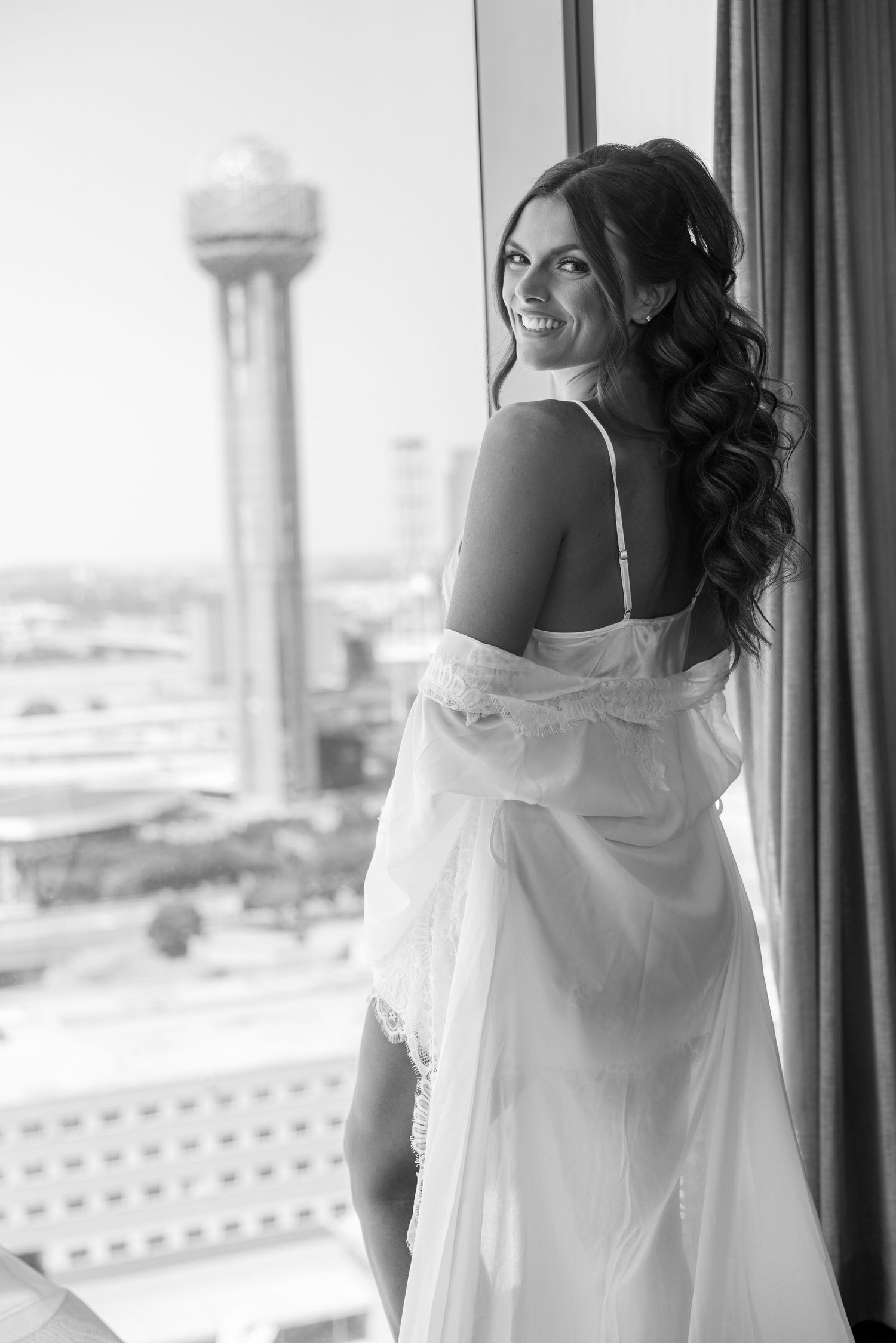 Black and white photo of a smiling bride in a white gown standing by a window with a city skyline view.