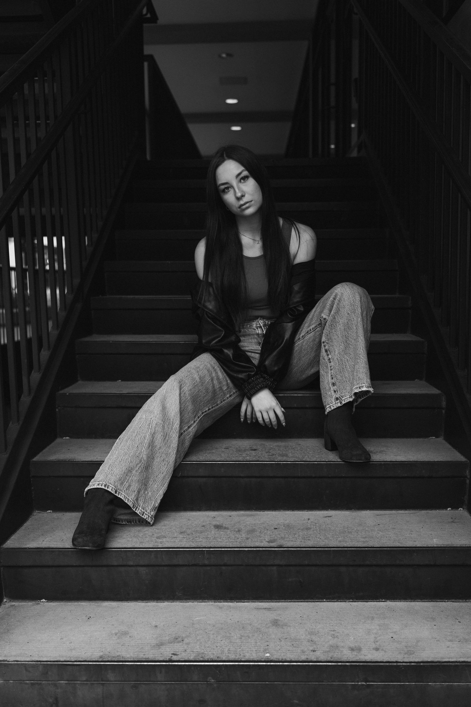 Young woman in black jacket and wide-leg jeans sitting on stairs in a moody black and white portrait photo.