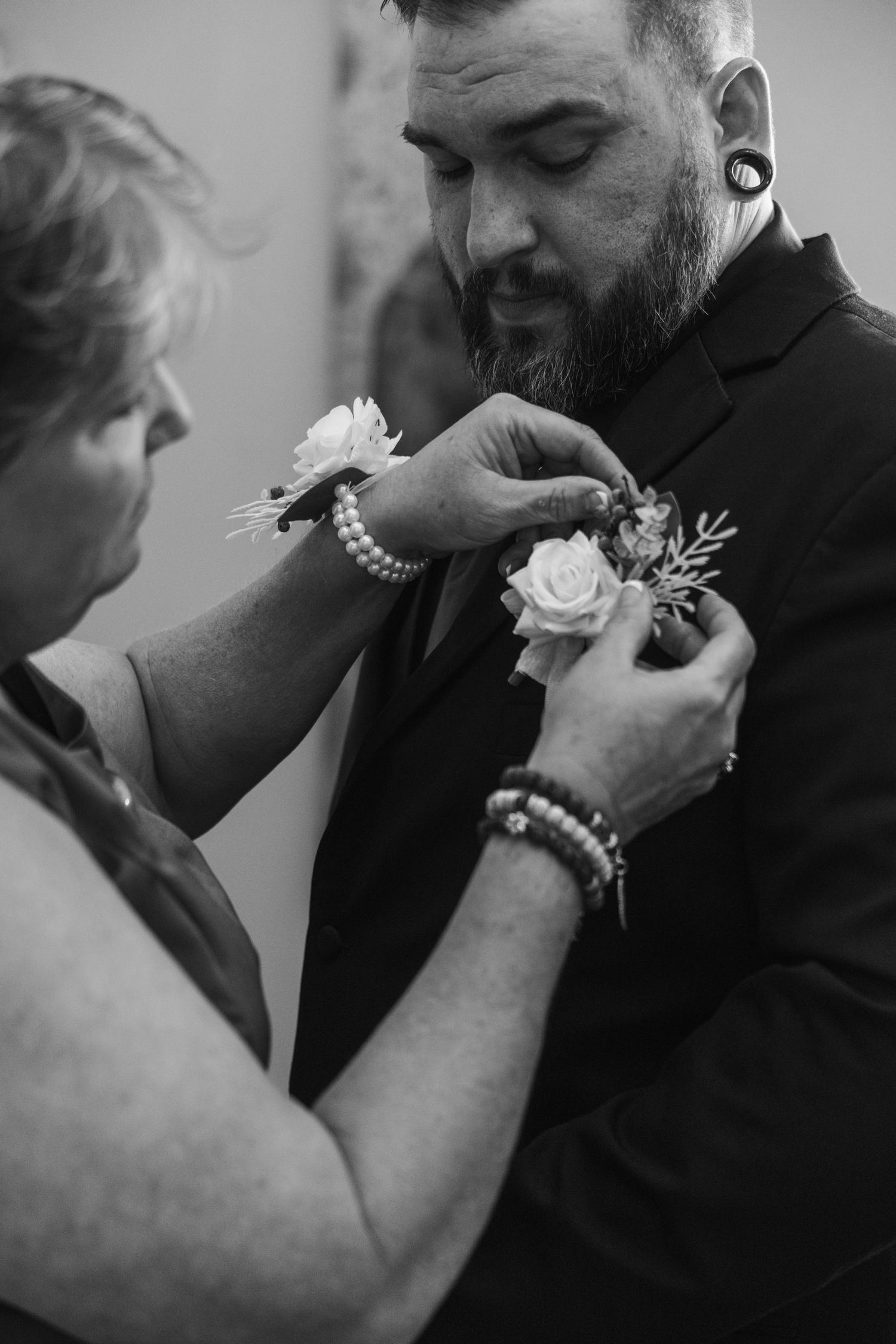 A woman pins a white rose boutonniere on a bearded groom in a black suit during wedding preparations.