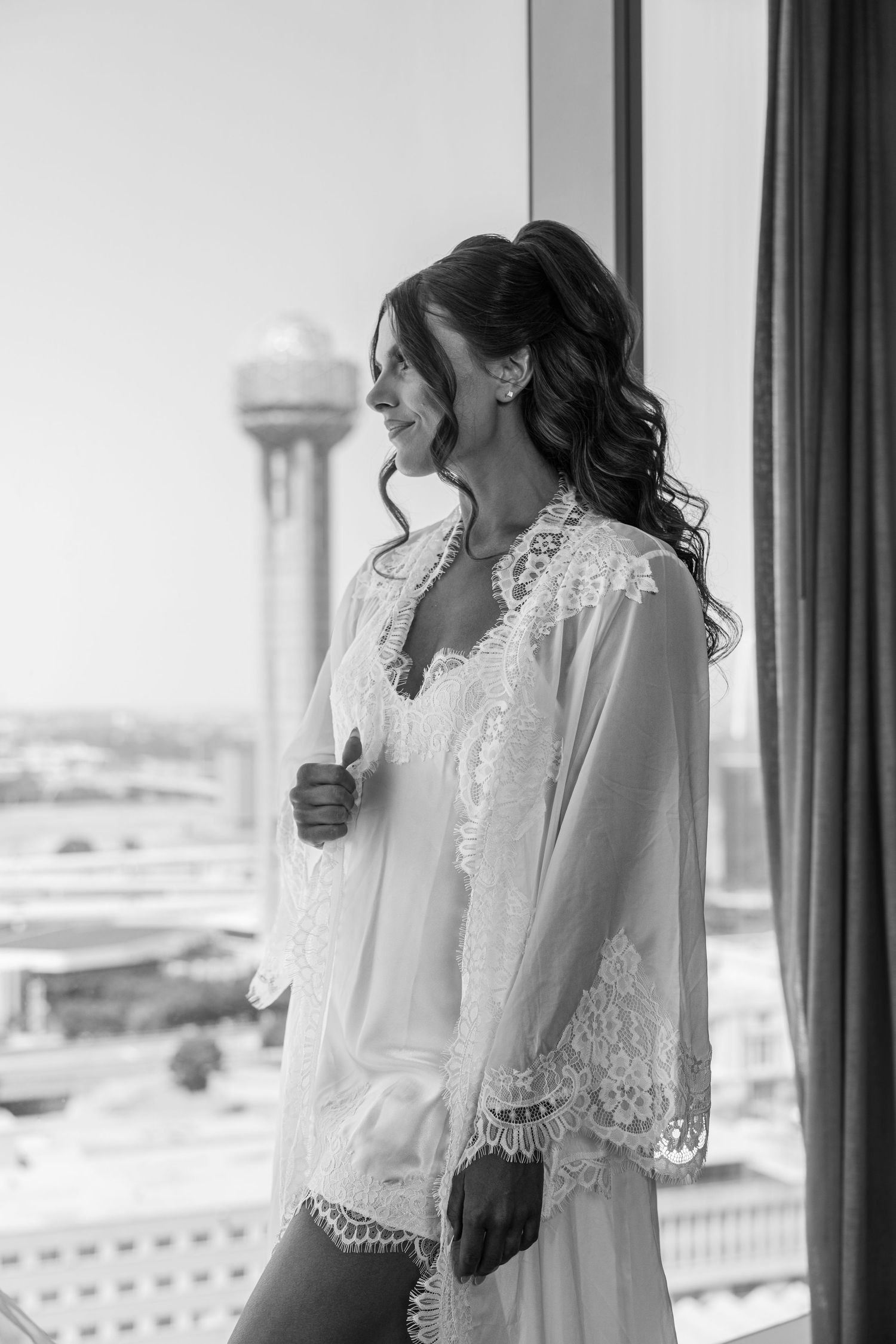 Black and white photo of a smiling bride in lace robe standing by a window with a city skyline view.