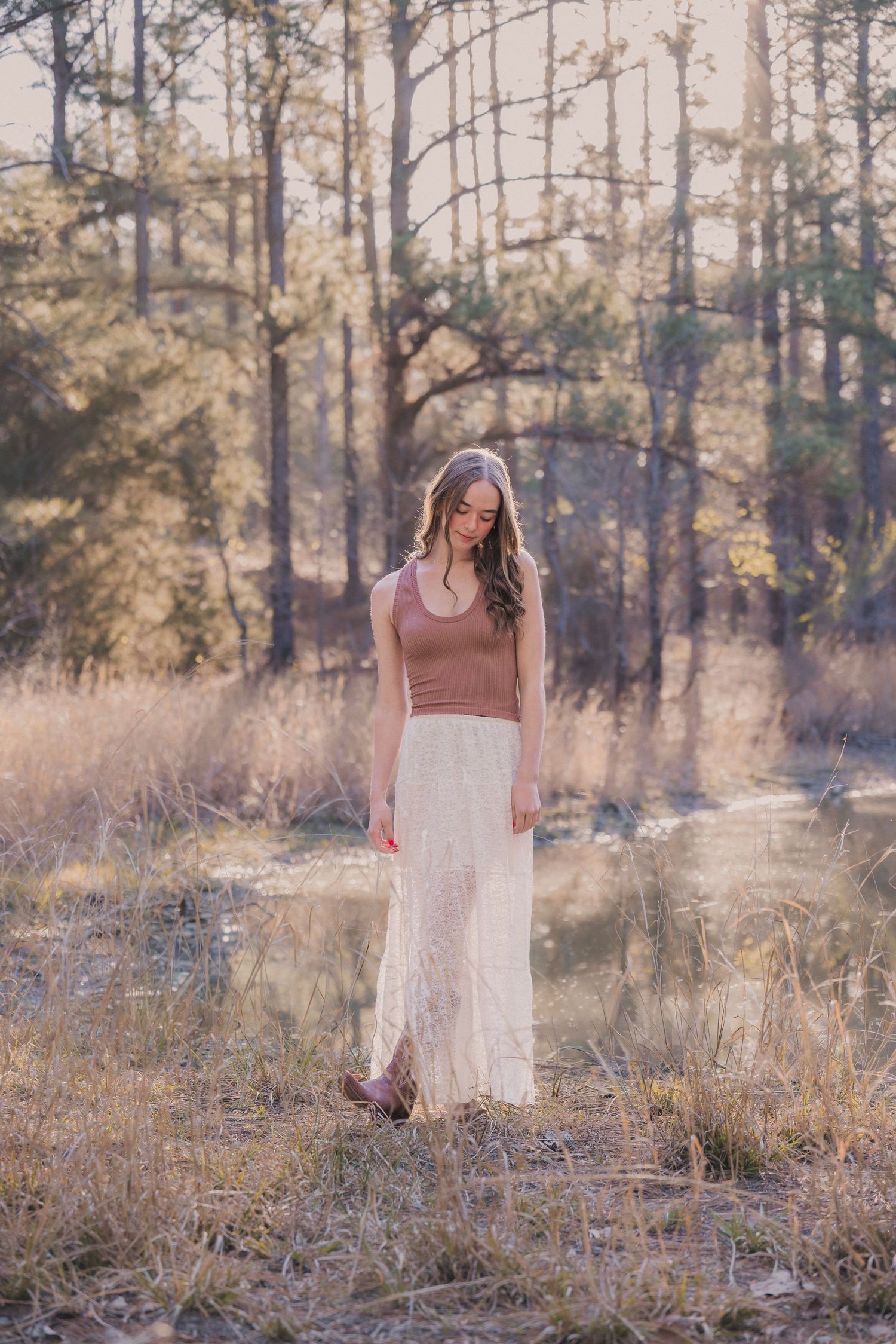 A dreamy forest scene with a white lace dress and mauve top against a backdrop of sunlit pine trees and tall grass.