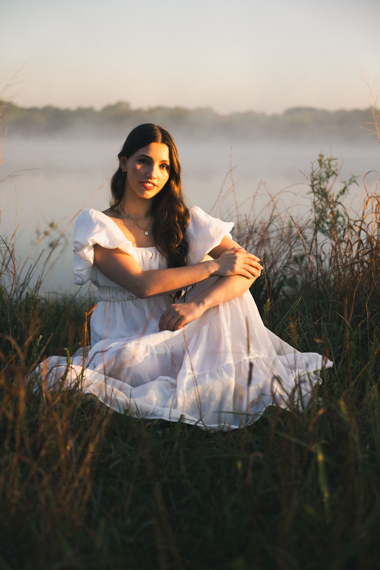 Young woman in white dress sitting in golden hour light by misty lake surrounded by tall grass.