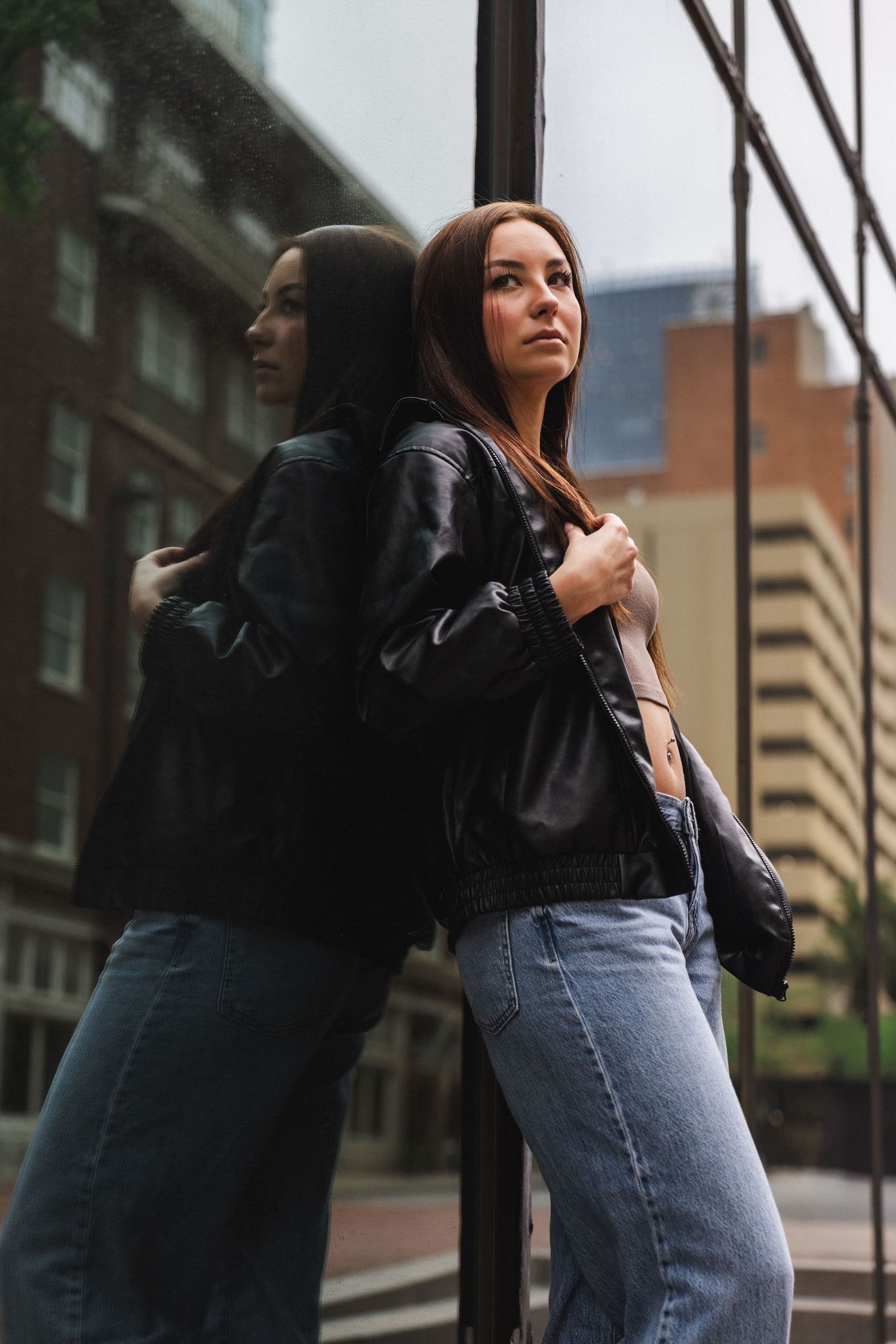 Young woman in black leather jacket and jeans leaning against reflective glass wall on urban street.