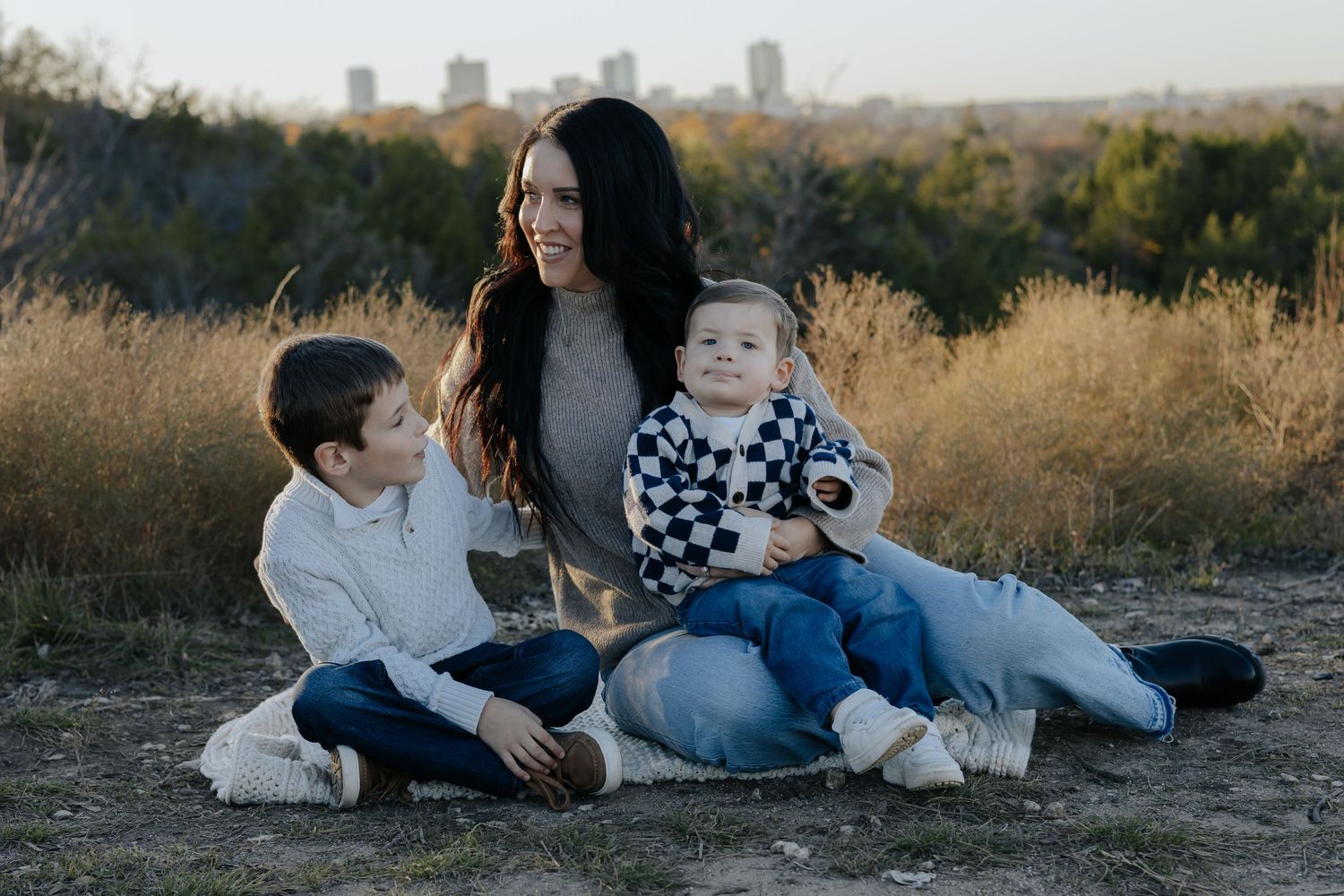 A family sits together in a grassy field during golden hour with a city skyline visible in the background.
