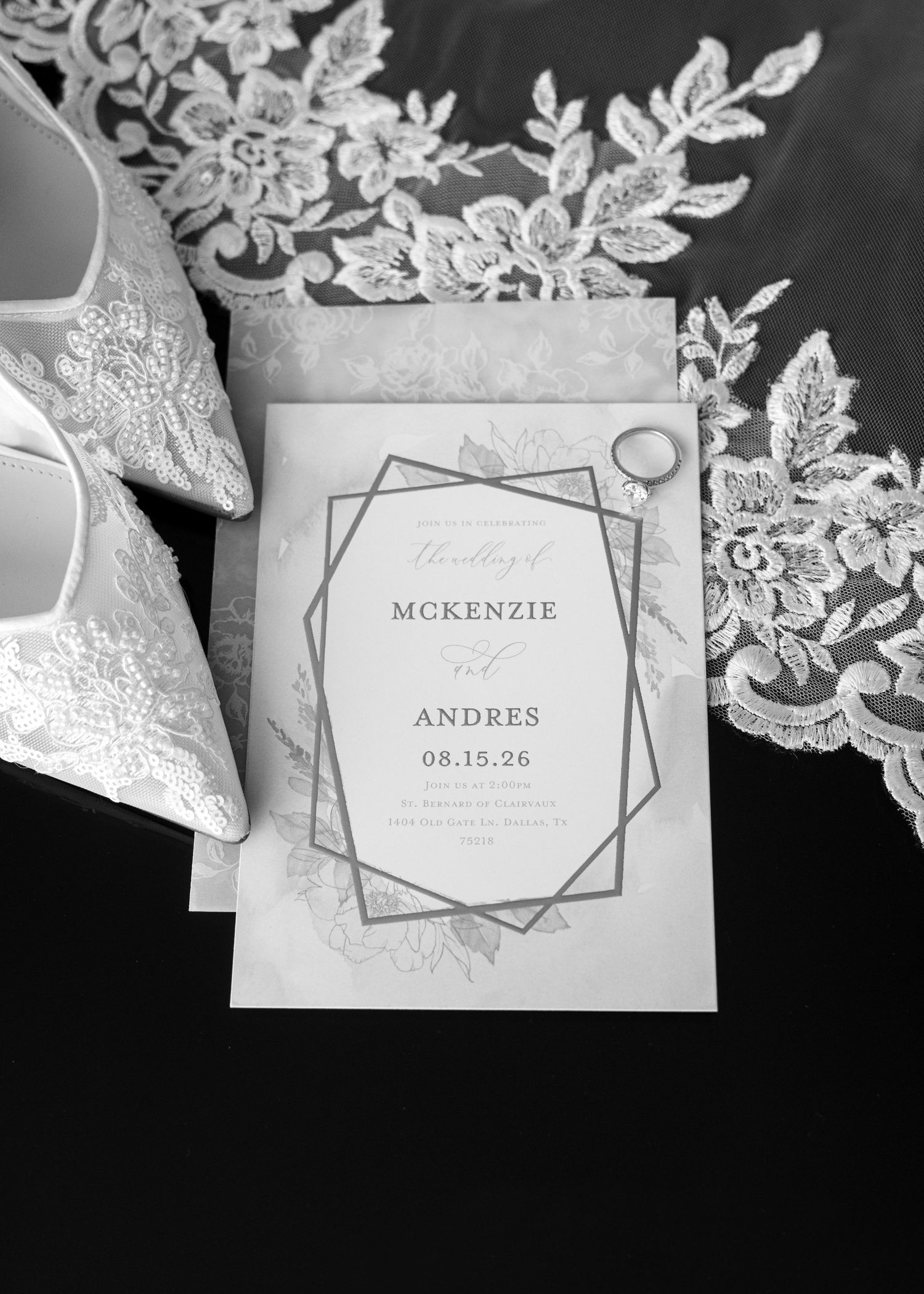 Black and white flat lay of wedding invitation for McKenzie and Andres, lace bridal shoes, veil, and ring.