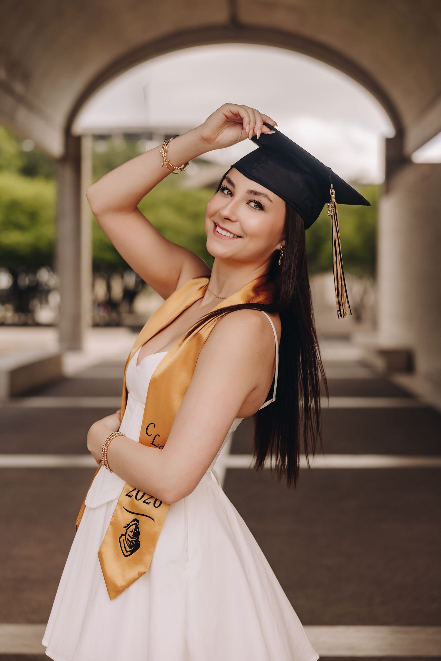 Smiling young woman in white dress and graduation cap holding tassel, wearing gold 2020 stole, posing under arched columns.