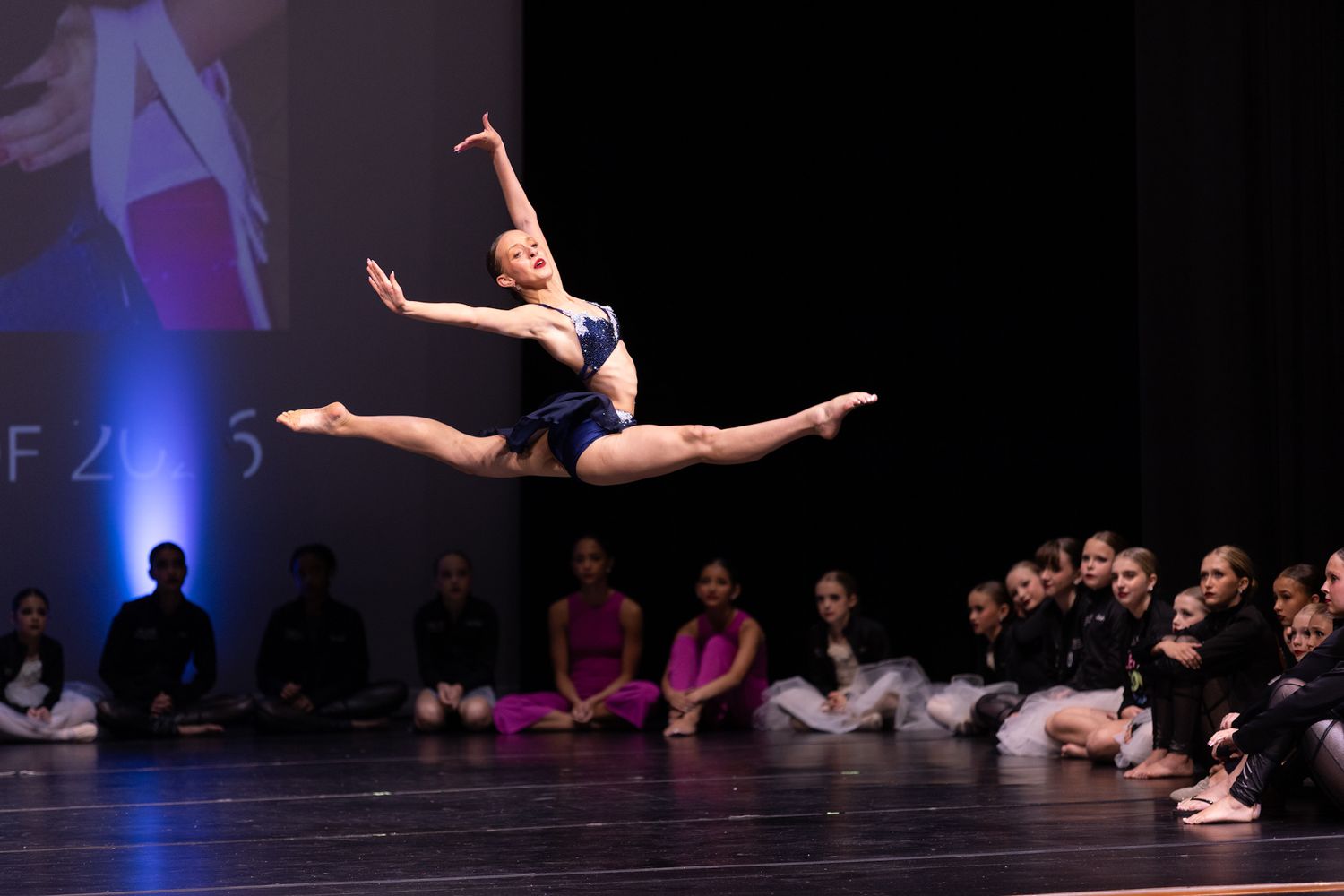 A ballet dancer performs a stunning grand jeté leap on stage during a dance competition, with an audience watching in the background.