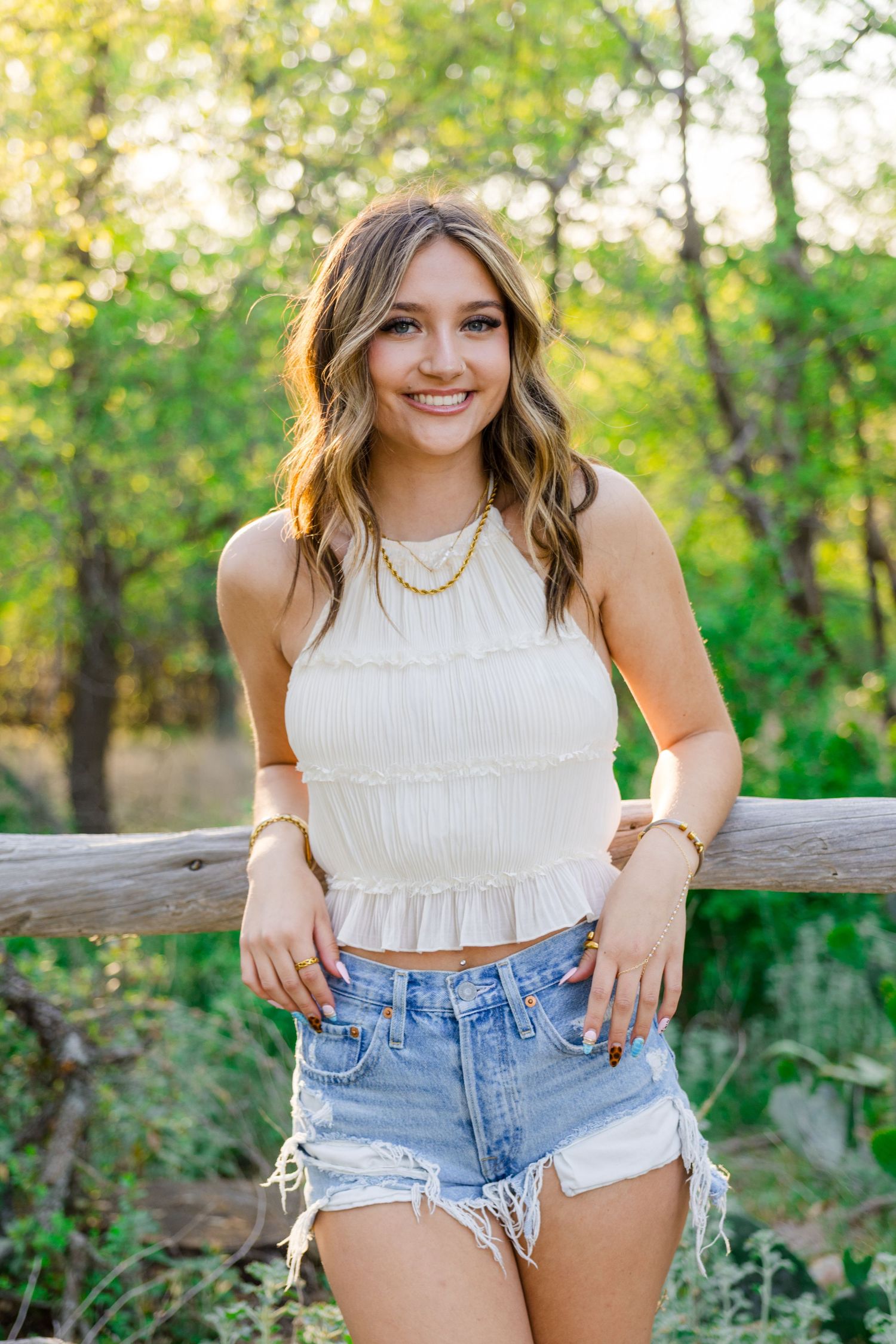 Young woman smiling in white crop top and denim shorts, leaning on wooden fence with green trees background.