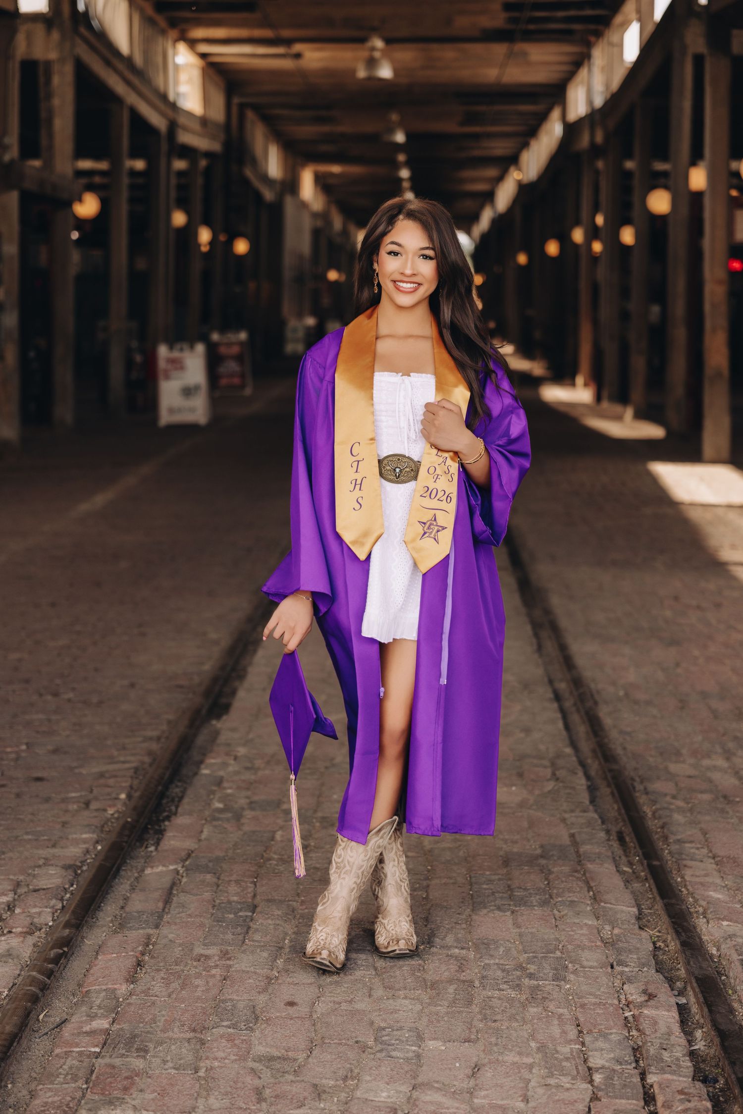 Young woman in purple graduation gown and gold stole holding cap, wearing cowboy boots in rustic hall.