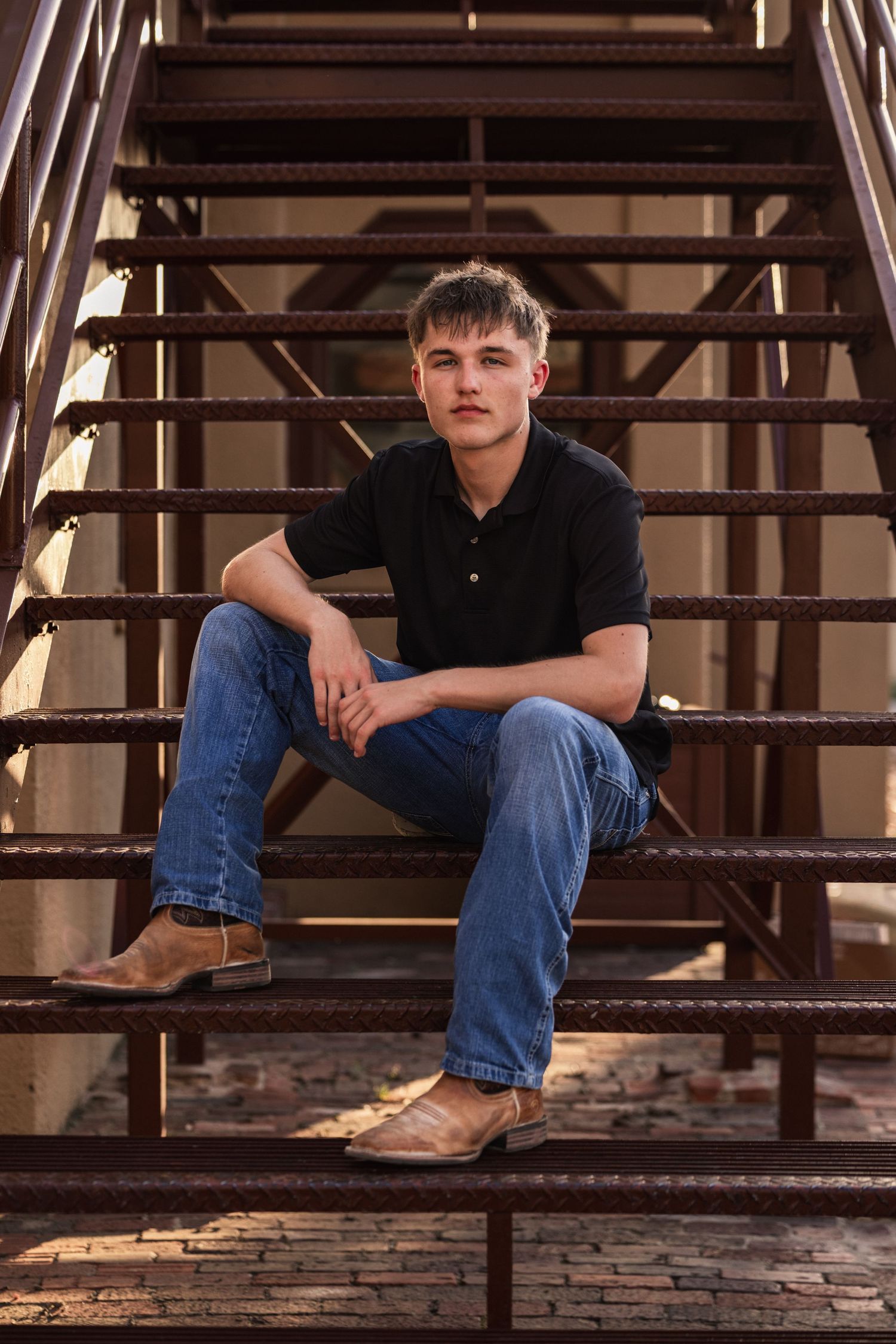 Young man in black polo and jeans sitting on rustic metal stairs, wearing tan cowboy boots in warm golden light.
