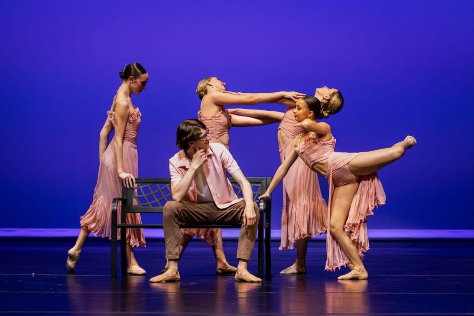Dancers in flowing beige costumes perform a contemporary dance piece on a stage illuminated by deep blue lighting.