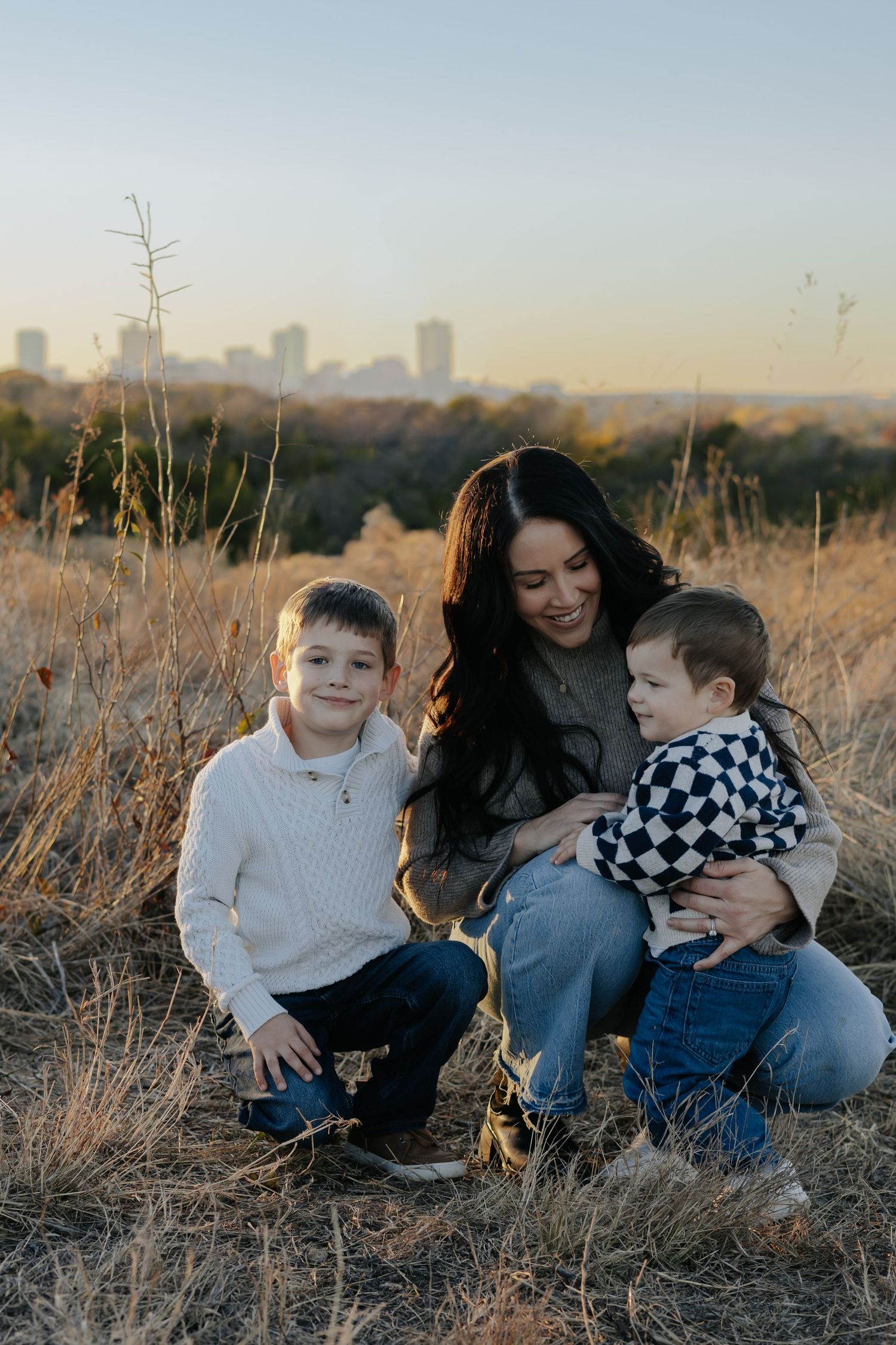 A family sits together in a grassy field at sunset with a city skyline visible in the background.
