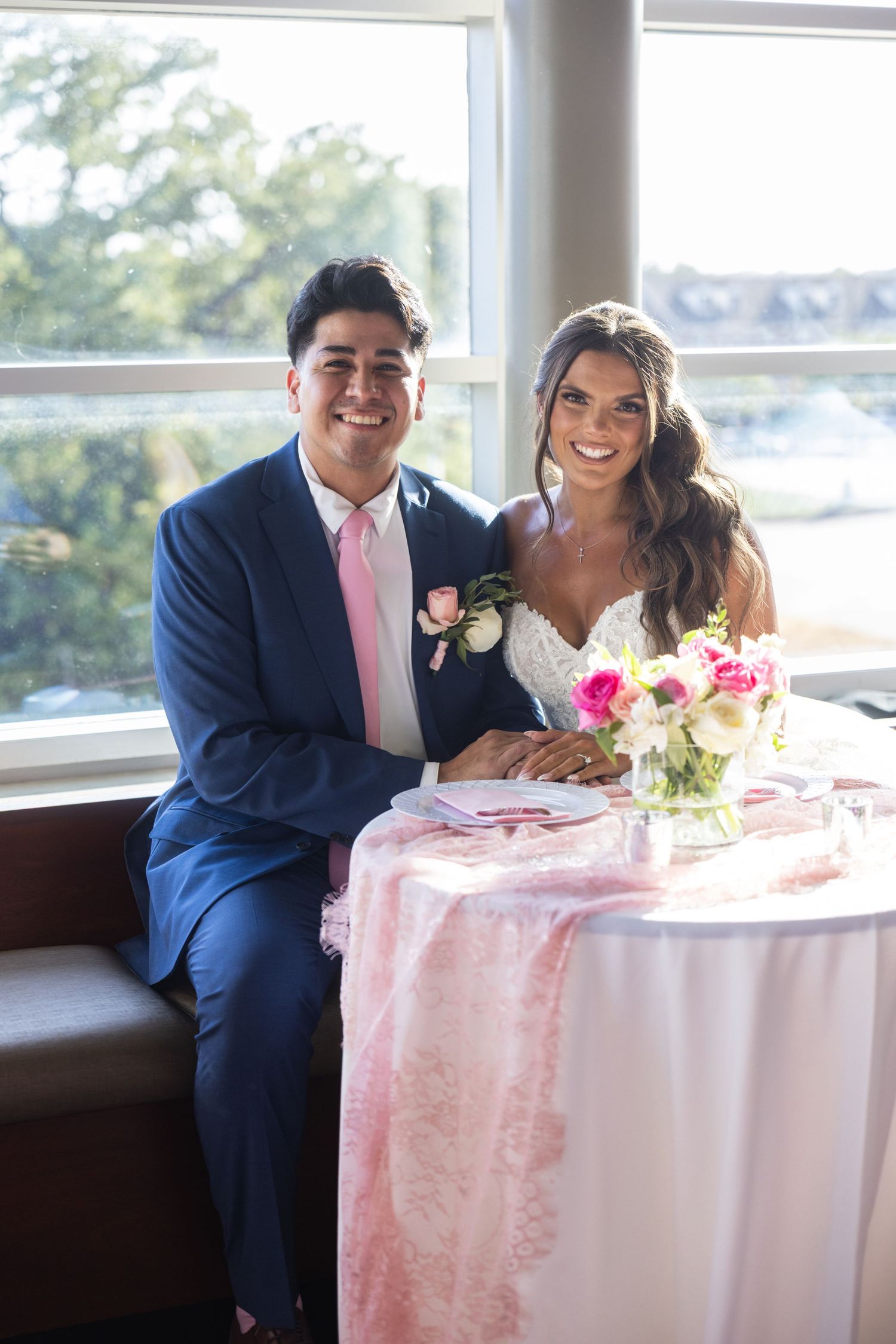 Smiling bride and groom seated at a pink floral sweetheart table by a window at their wedding reception.