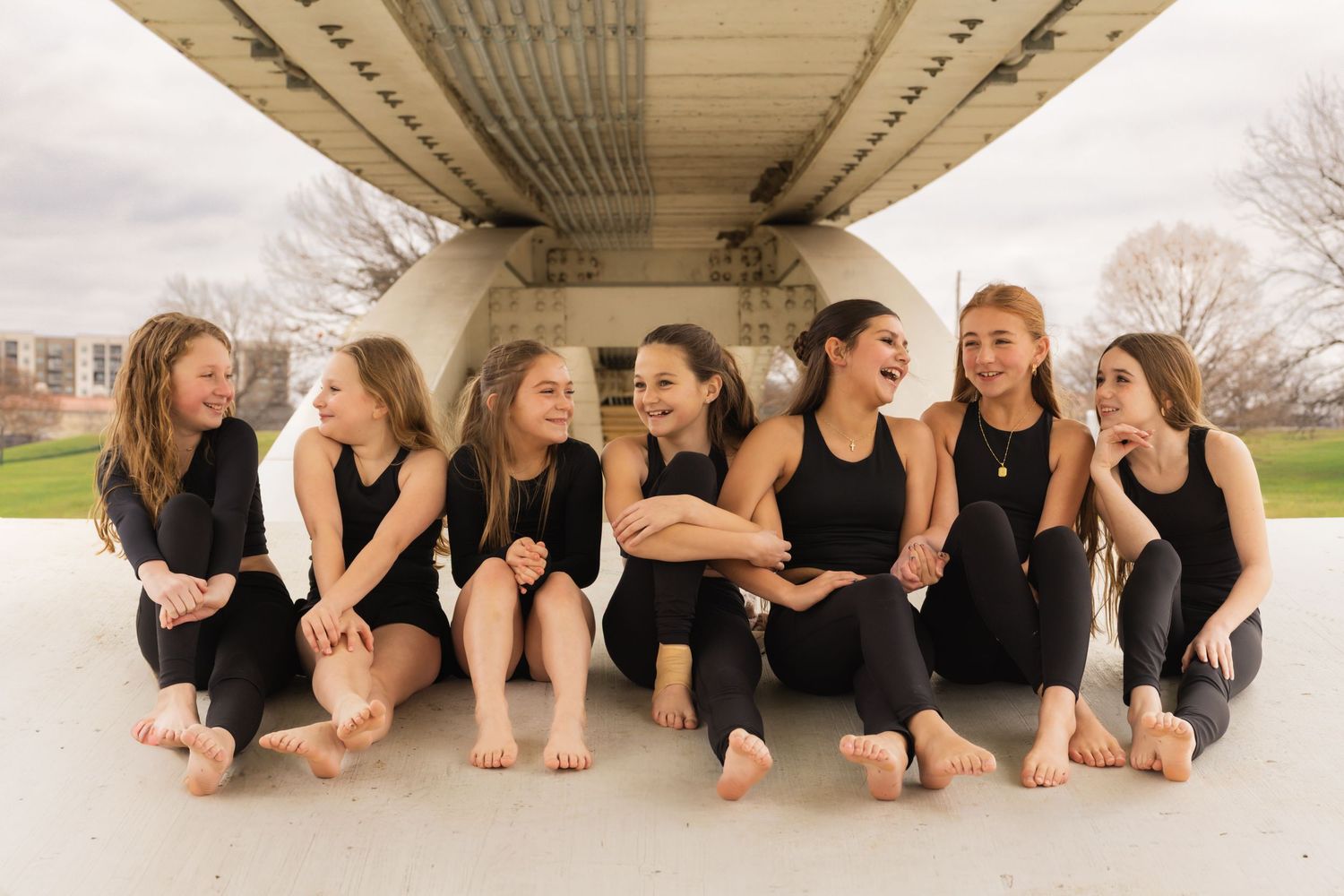 Group of dancers in black outfits lying on ground in artistic formation during outdoor photo shoot.