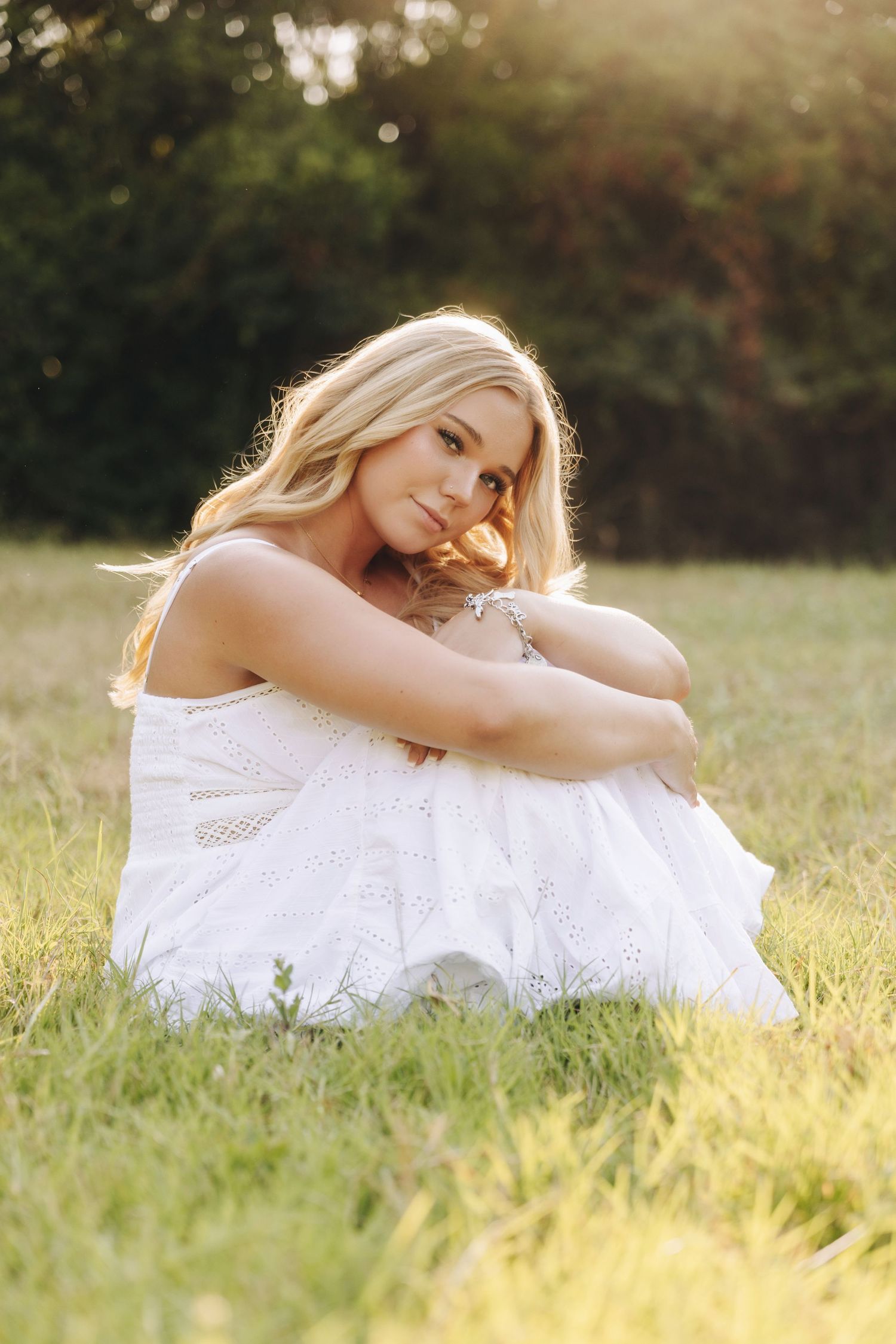 Blonde woman in white dress sitting on grass in golden sunlight, hugging her knees and smiling softly.