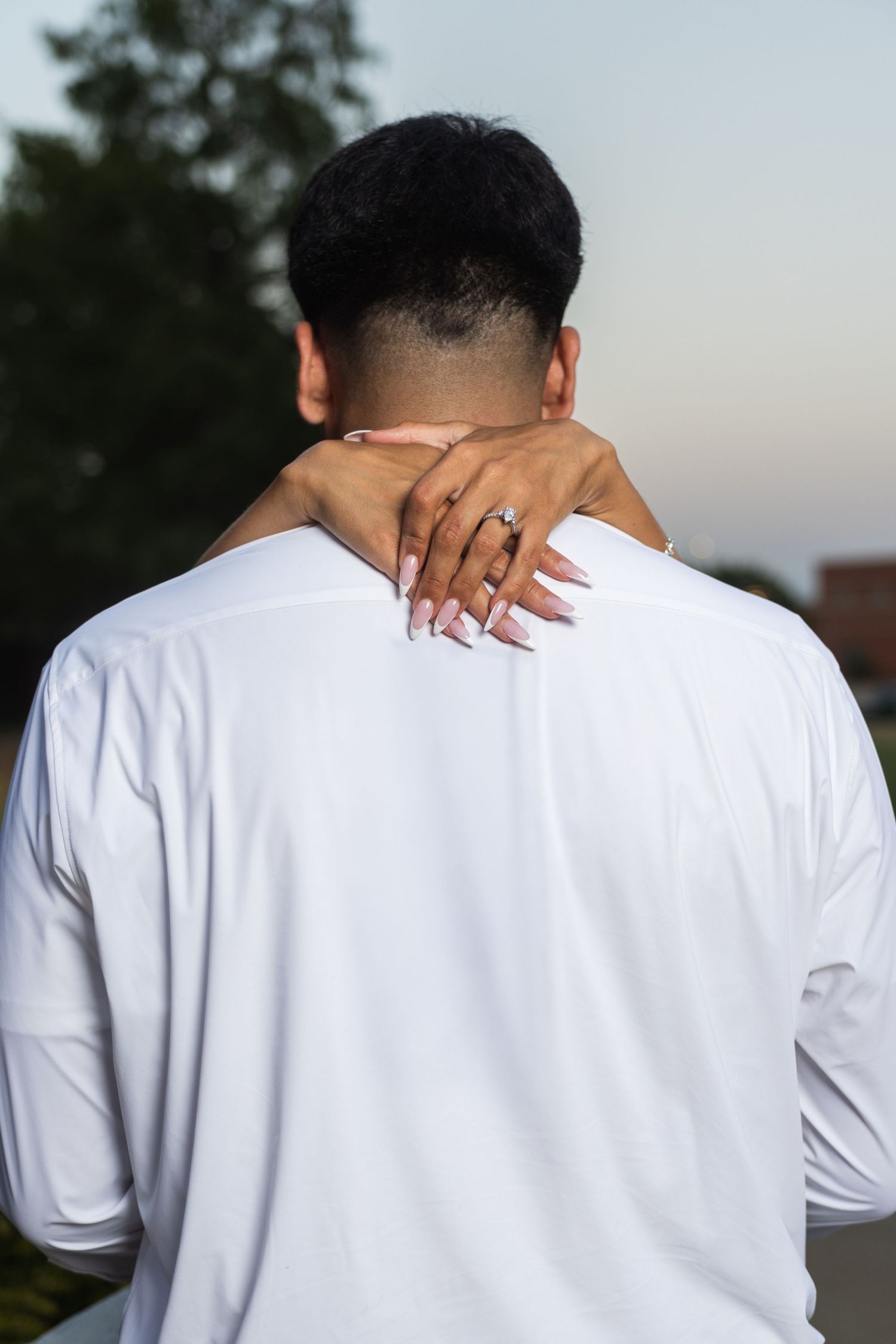 A woman with manicured nails and engagement ring embraces a man in white shirt from behind outdoors.