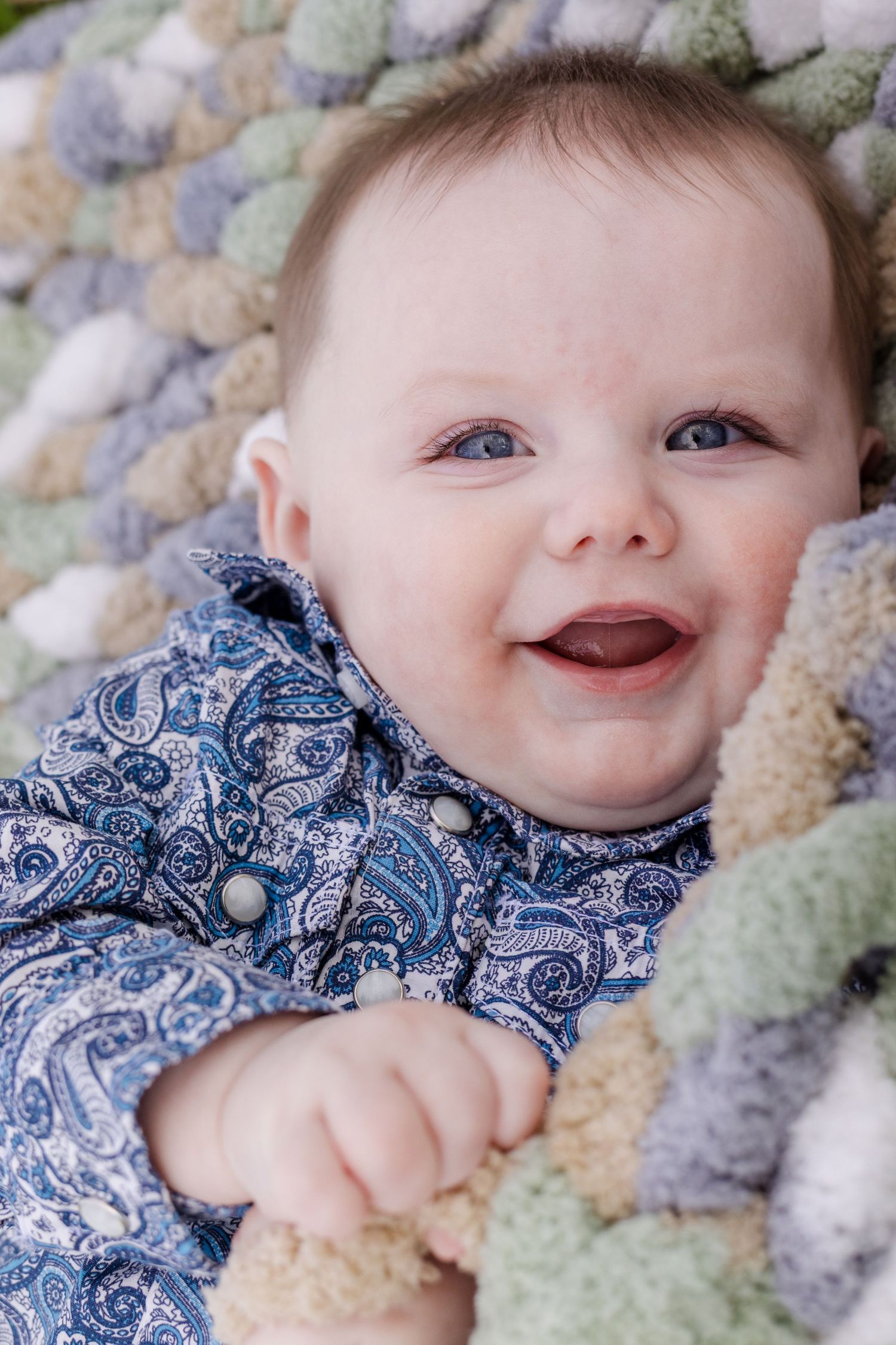 Smiling baby with blue eyes lying on a colorful chunky knit blanket wearing a blue paisley outfit.