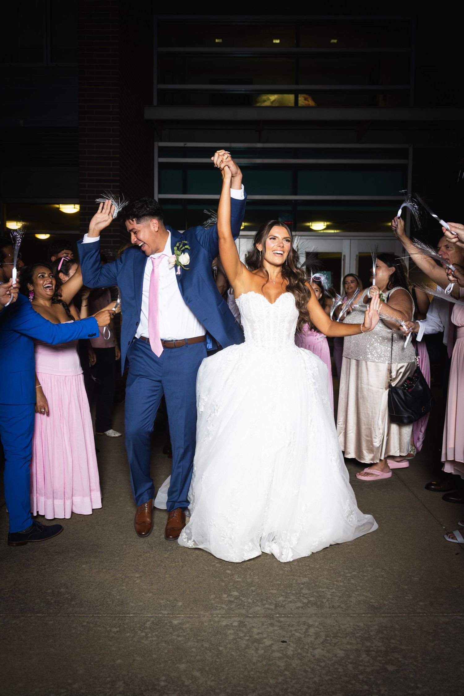 Bride and groom celebrate their wedding exit, raising hands joyfully while guests cheer outside at night.