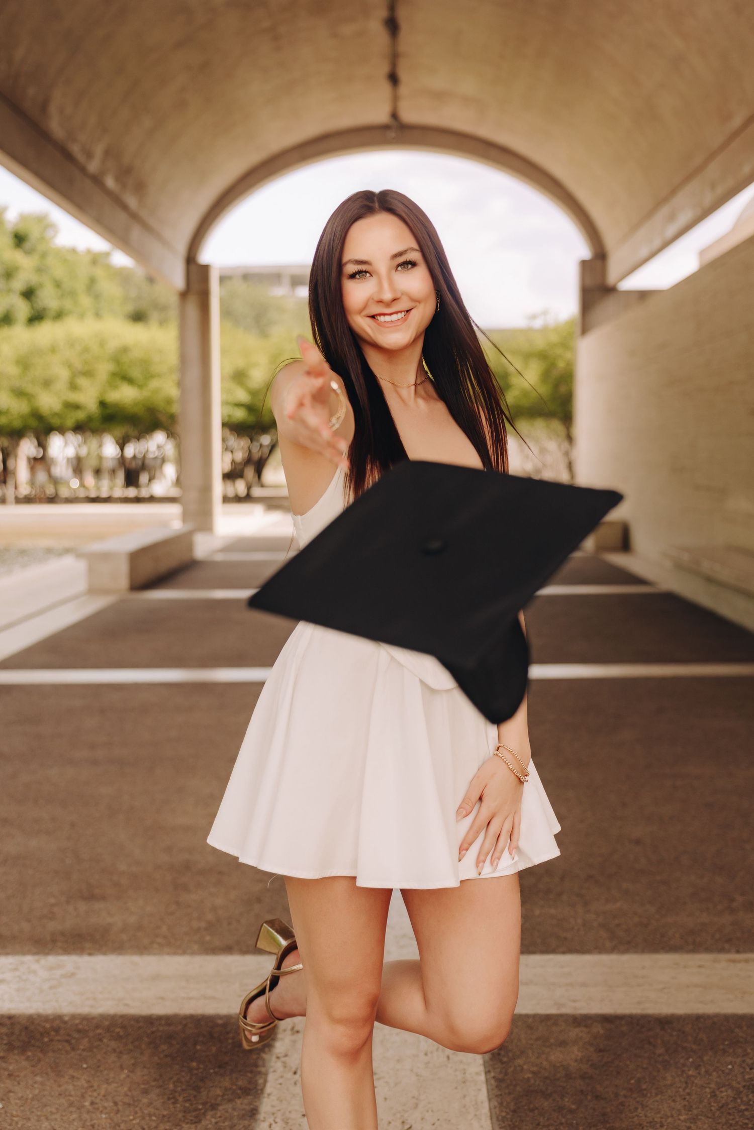 Smiling brunette graduate holding mortarboard cap in white skirt under arched walkway outdoors.