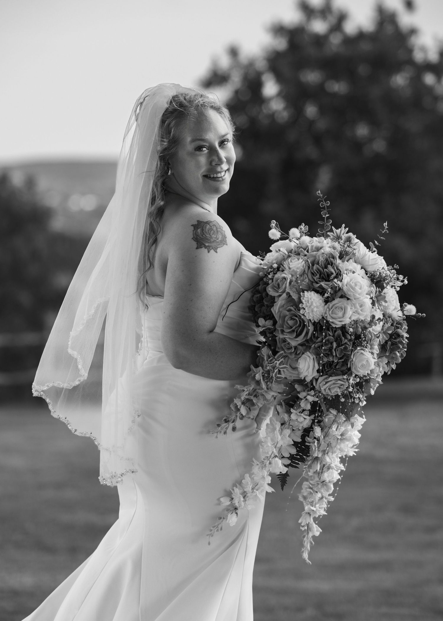 Bride in strapless gown and veil holds cascading floral bouquet, smiling over shoulder in black and white wedding portrait.