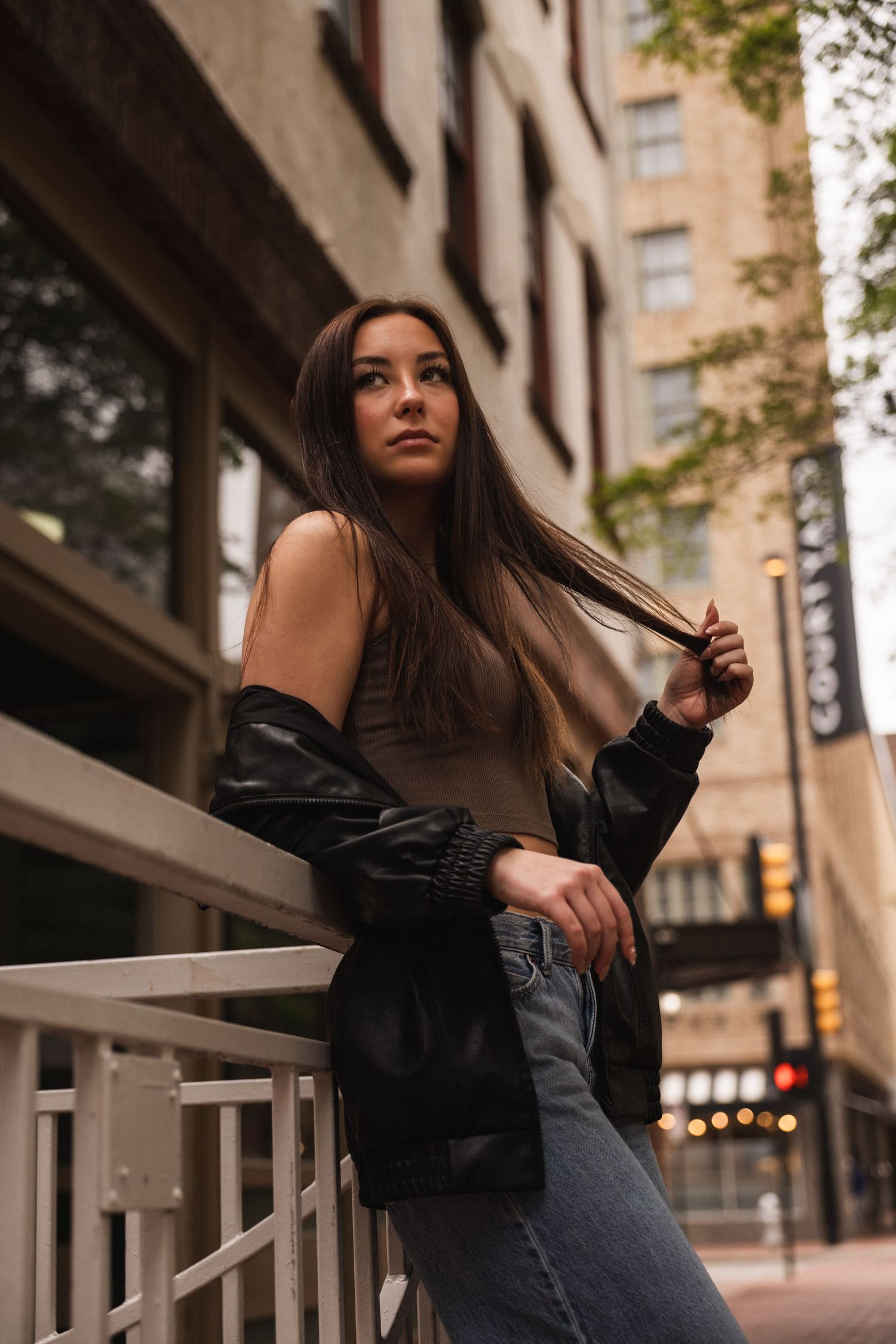 Young woman in black leather jacket and jeans leaning on urban railing with city buildings in background.