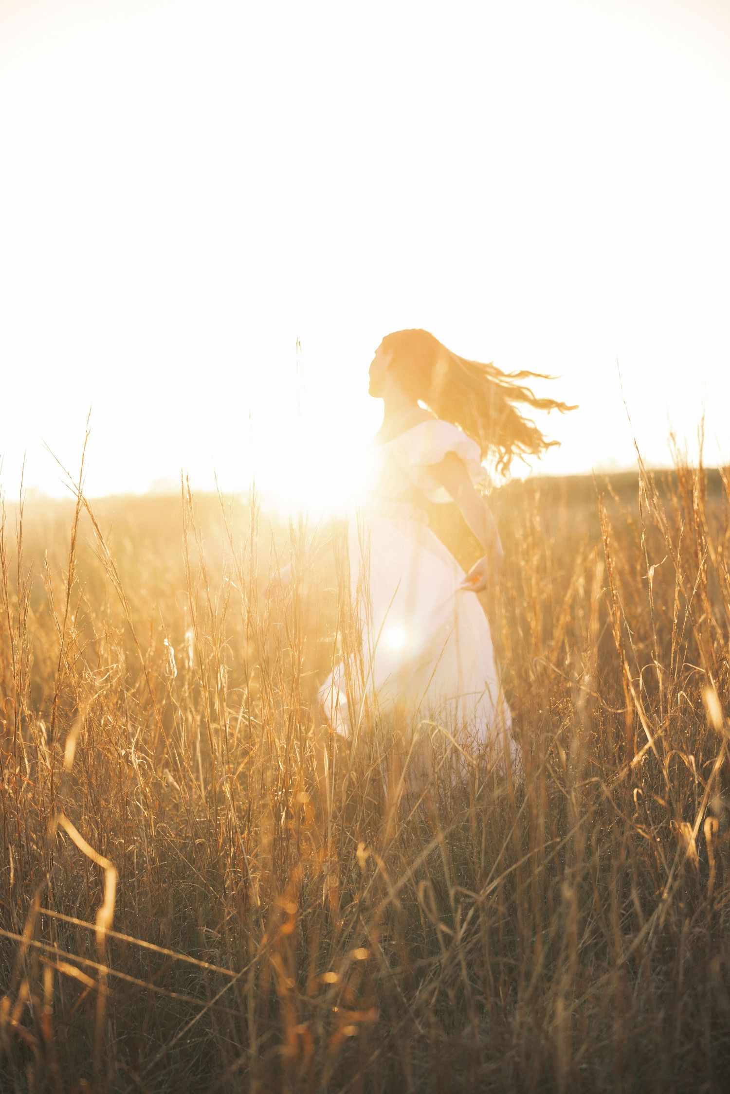 Woman in white dress standing in golden field at sunset, hair blowing in wind, backlit by warm sunlight.