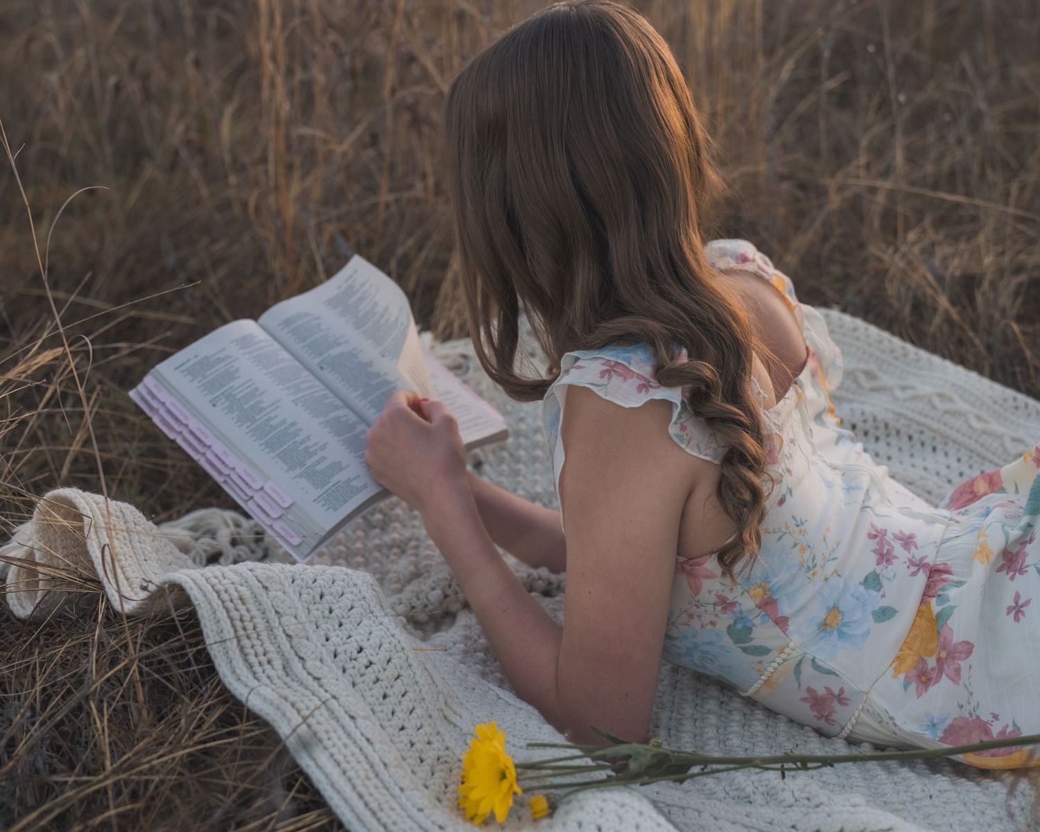 A person in a floral dress lies on a white blanket reading a book outdoors in nature.