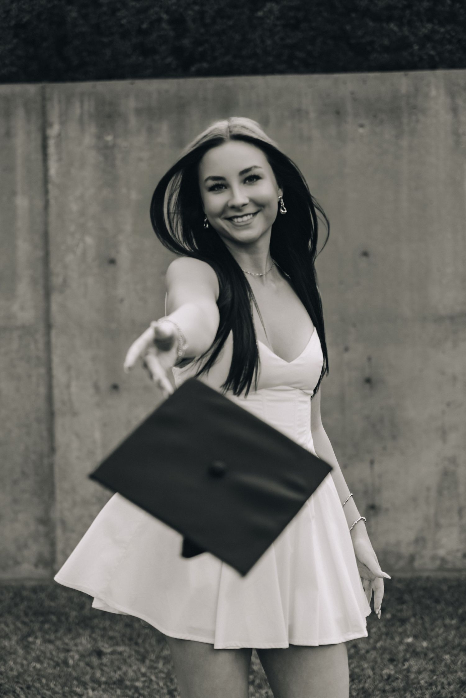 Smiling young woman in white dress tossing graduation cap outdoors in black and white photo.