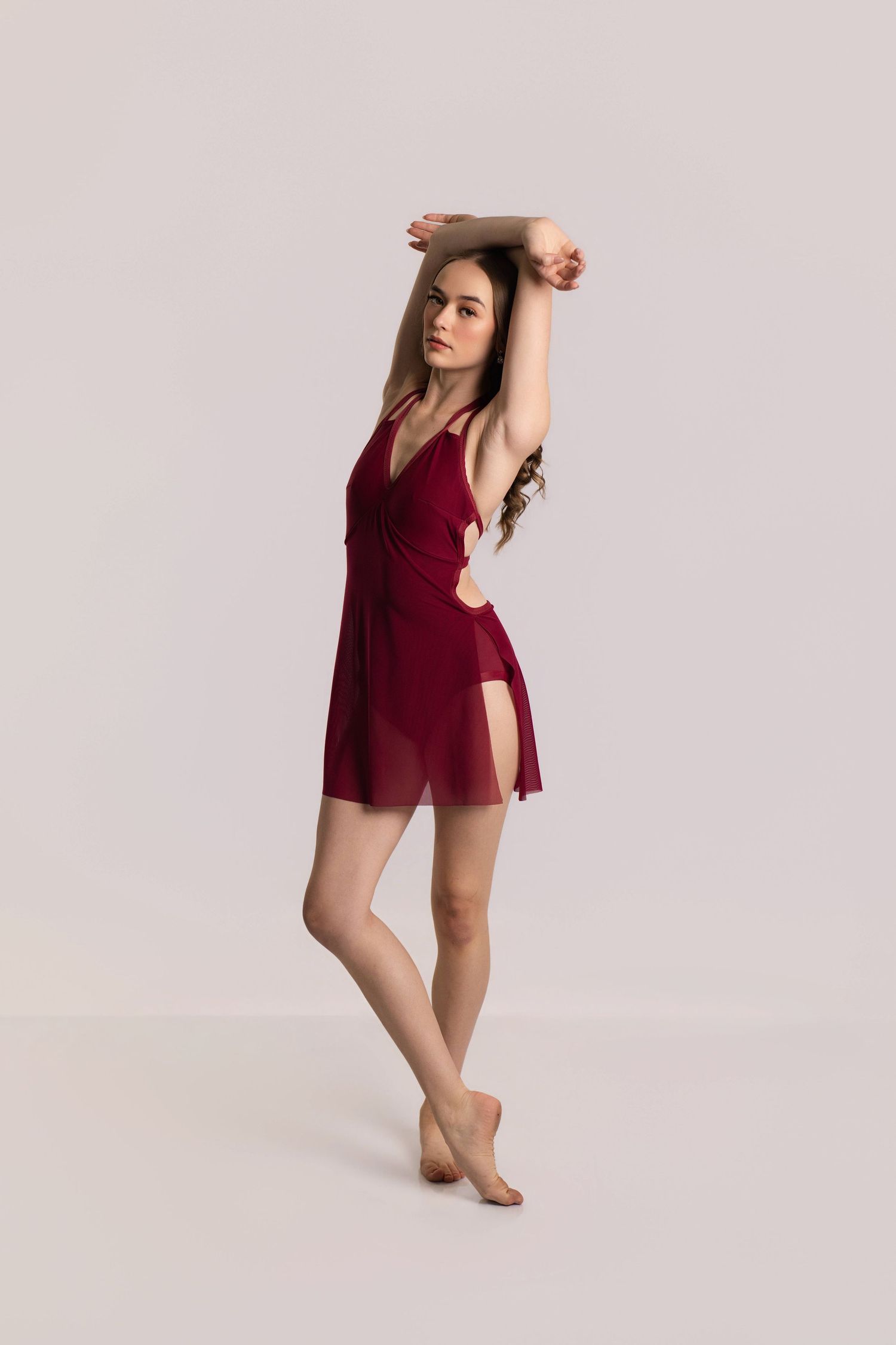 Young woman in a dark red wrap dress poses gracefully with arms raised overhead against a light gray background.