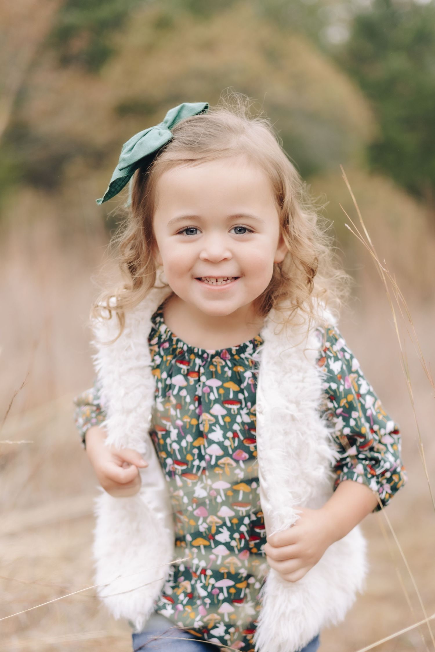 A child wearing a floral dress and white furry vest smiles while standing in a natural outdoor setting.