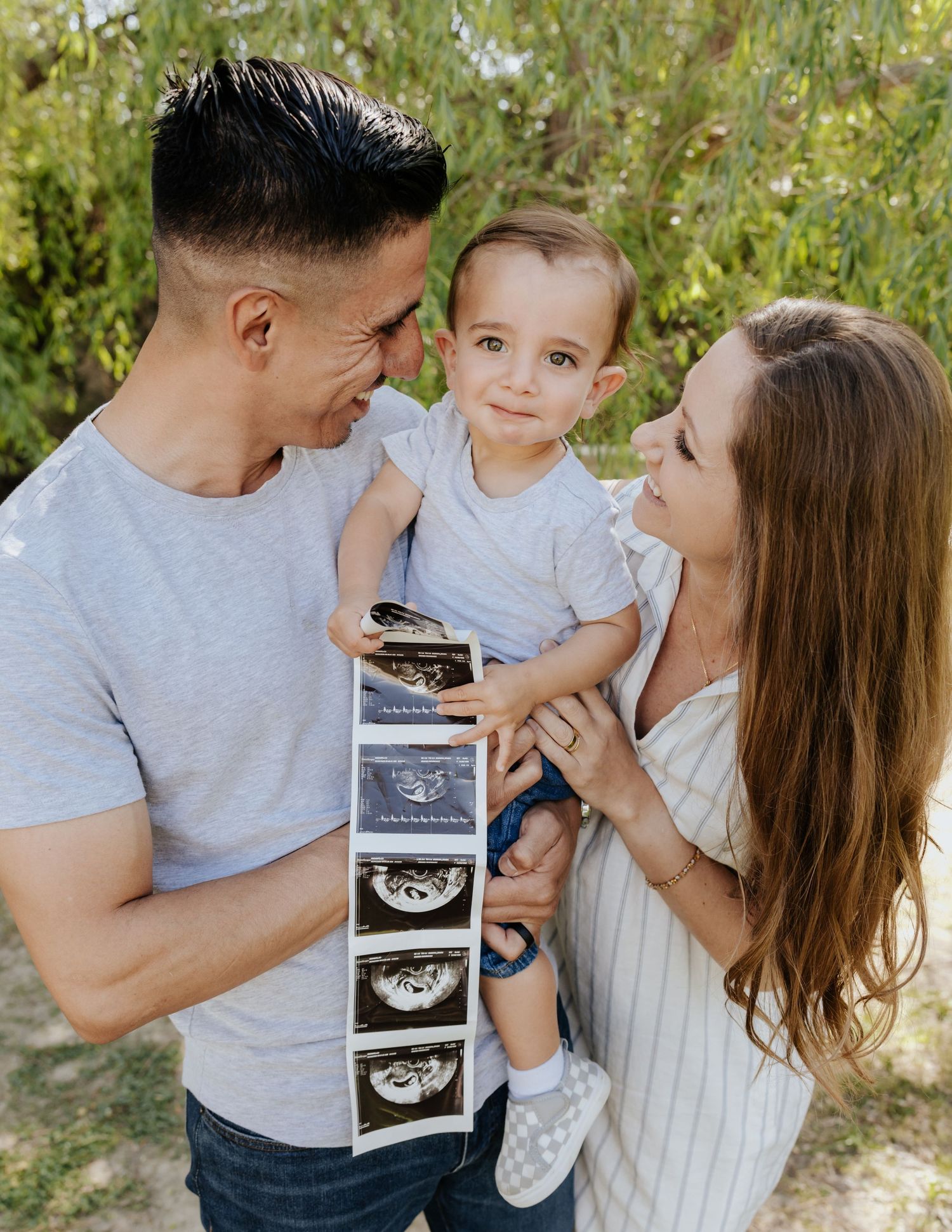 A couple holding their toddler who displays ultrasound photos, announcing a second pregnancy outdoors.