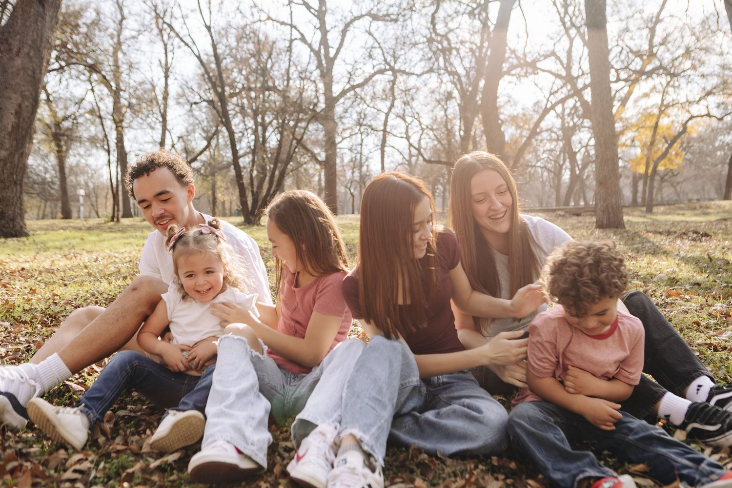 A group sits together laughing and having fun on fallen autumn leaves in a park during golden hour.
