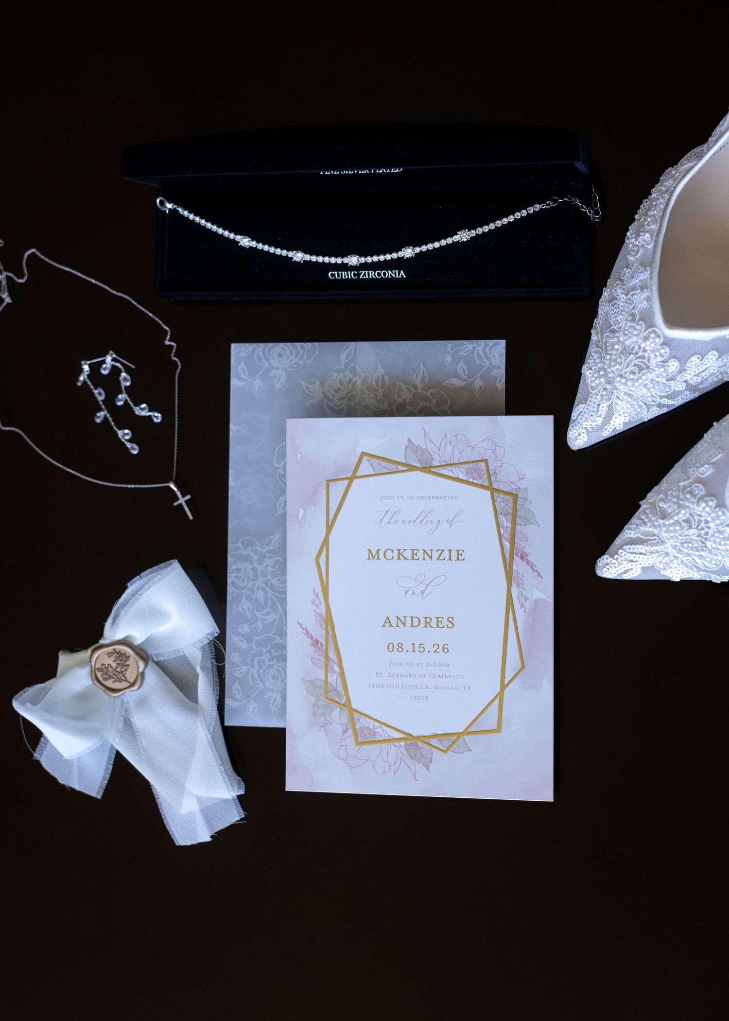 Elegant wedding flat lay featuring gold-framed invitation for McKenzie and Andres, diamond necklace, lace heels, and white ribbon bouquet.