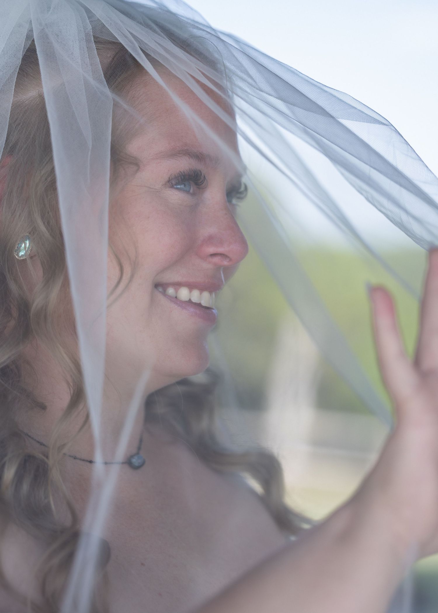 Smiling bride under a sheer white veil, wearing a necklace, with soft natural light in the background.