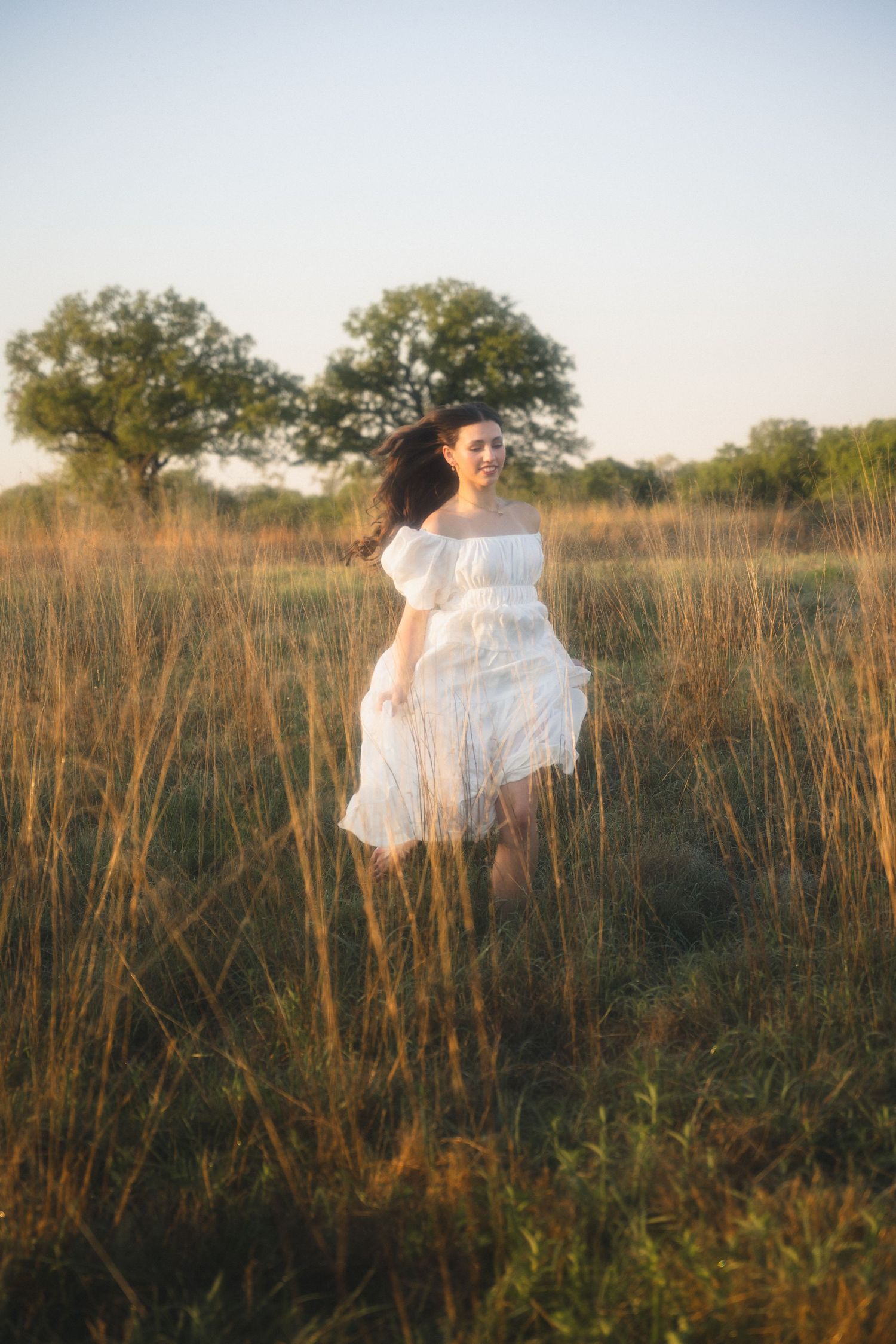 Young woman in white off-shoulder dress running through golden field at sunset with trees in background.