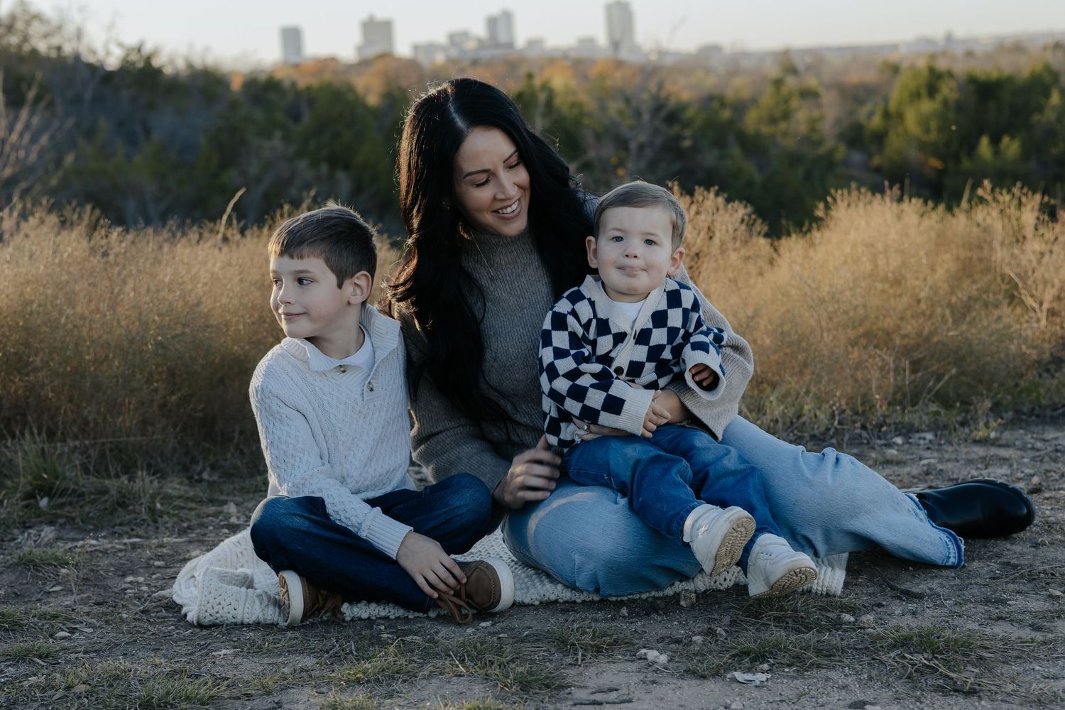 Family relaxing together on grassy hillside with city skyline in background during golden hour.