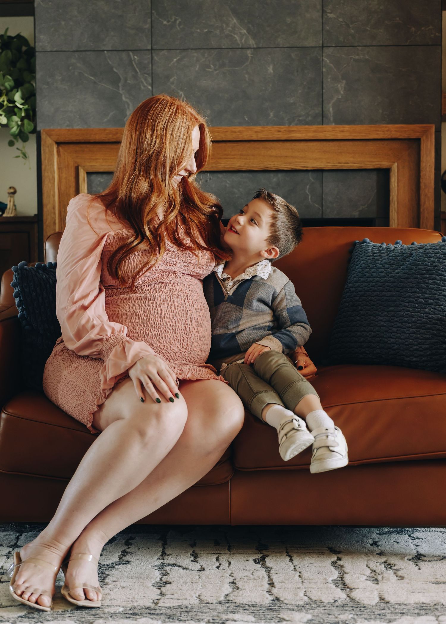 A pregnant woman in a pink dress sits on a brown leather couch with a young child, sharing an intimate moment at home.