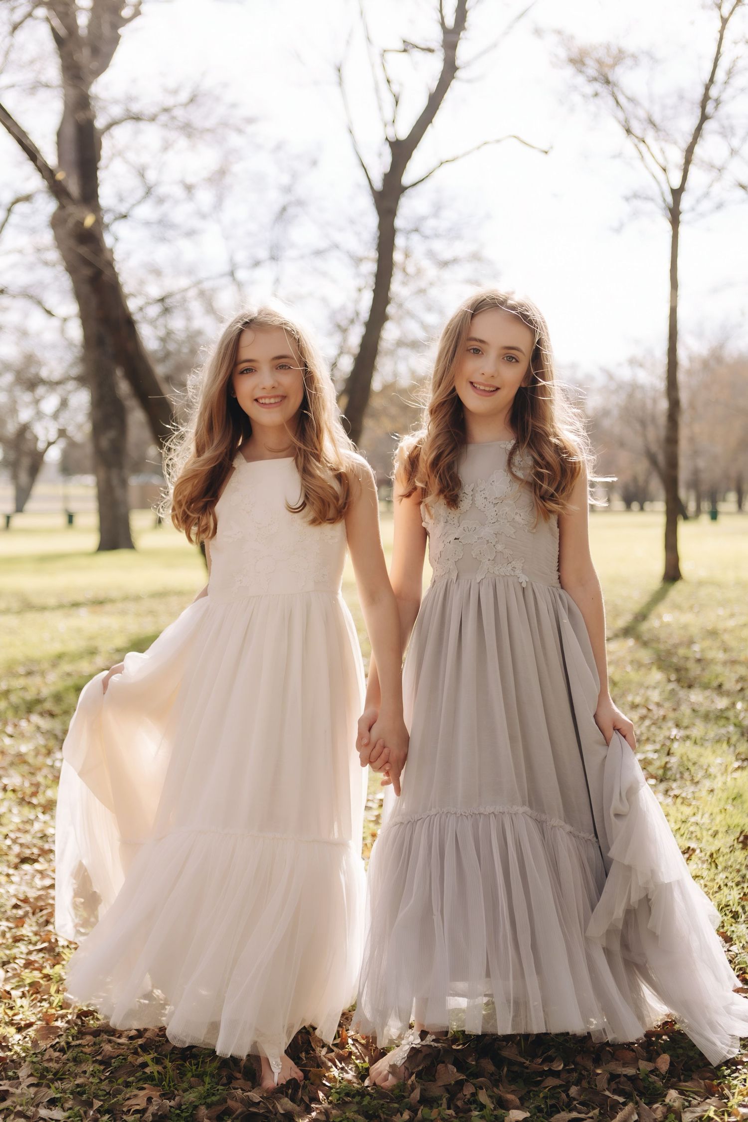 Two young girls in flowing white and gray dresses run and play together in a sunlit park setting.