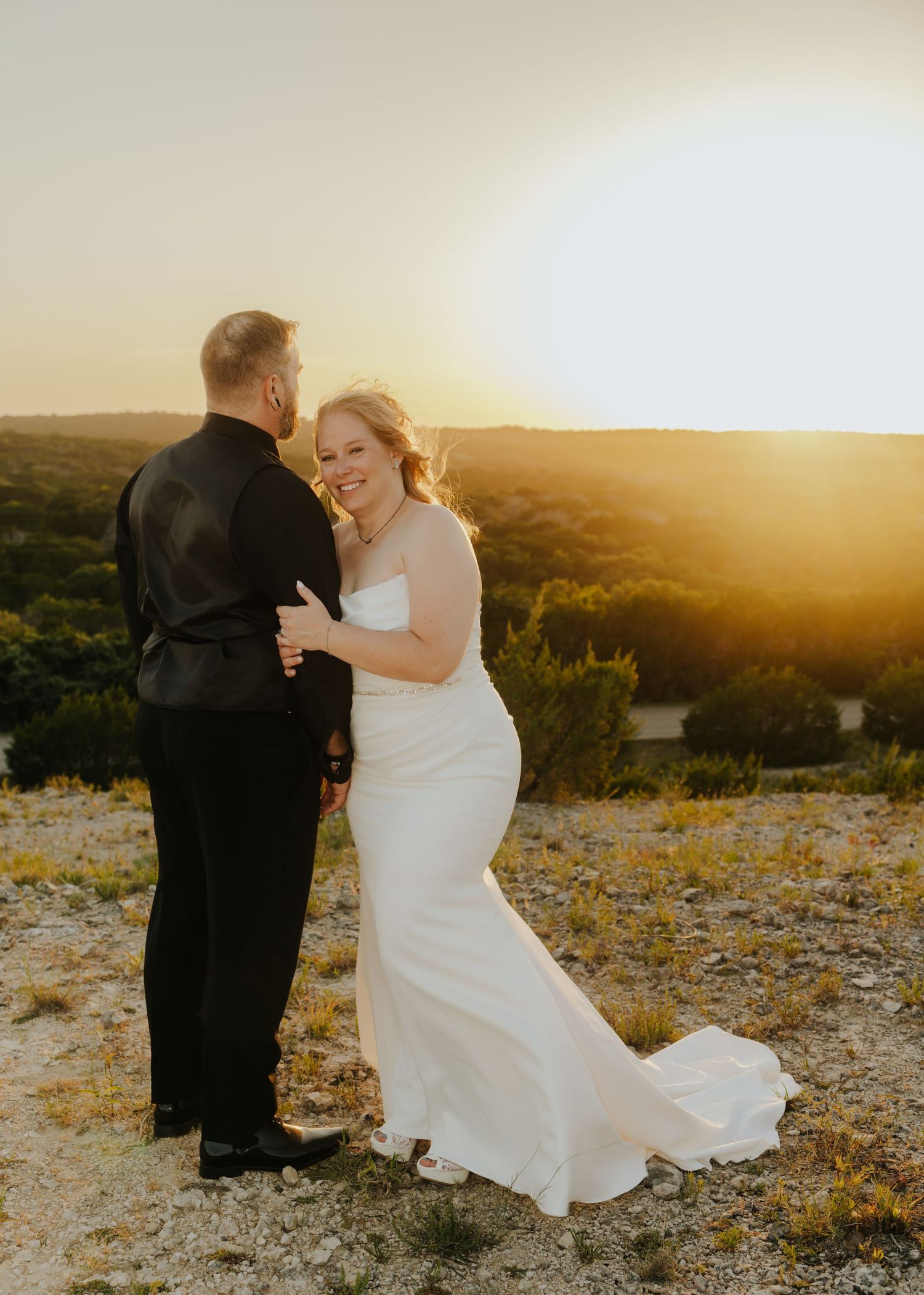 Bride and groom dancing at golden sunset on scenic hilltop, bride in elegant white gown smiling.