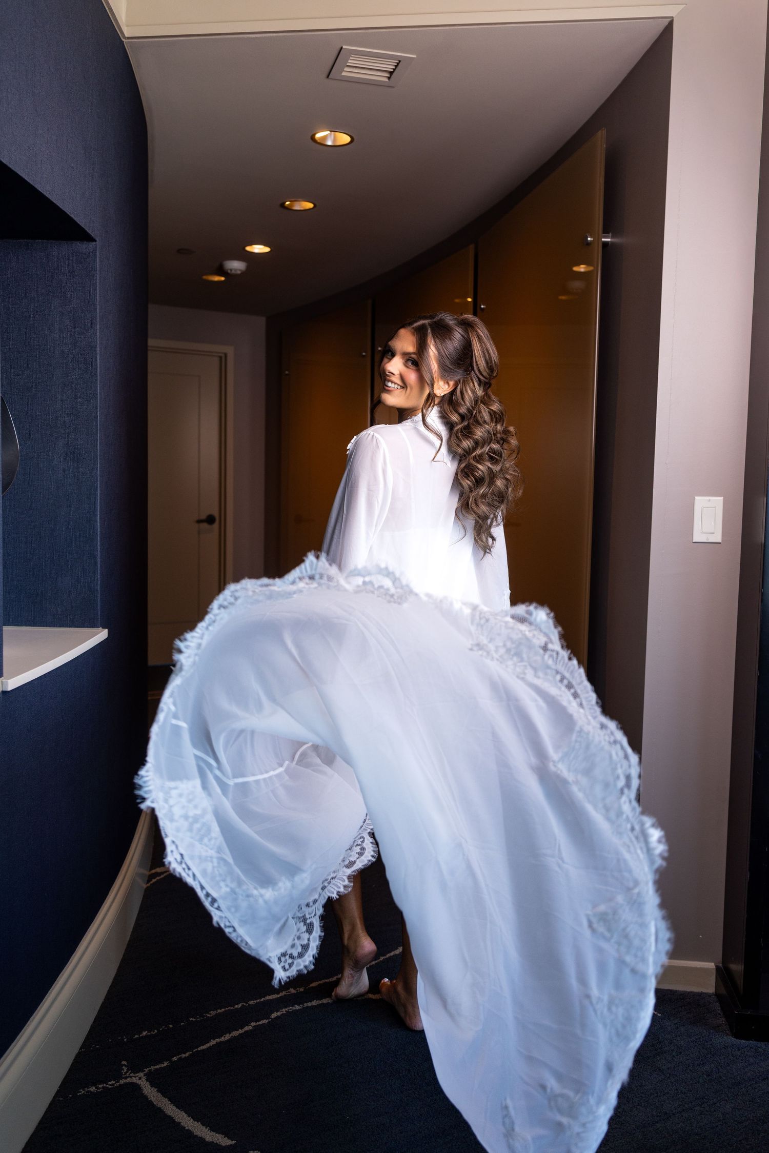 Bride in flowing white wedding dress twirling in hotel hallway, smiling over shoulder with elegant lace gown.